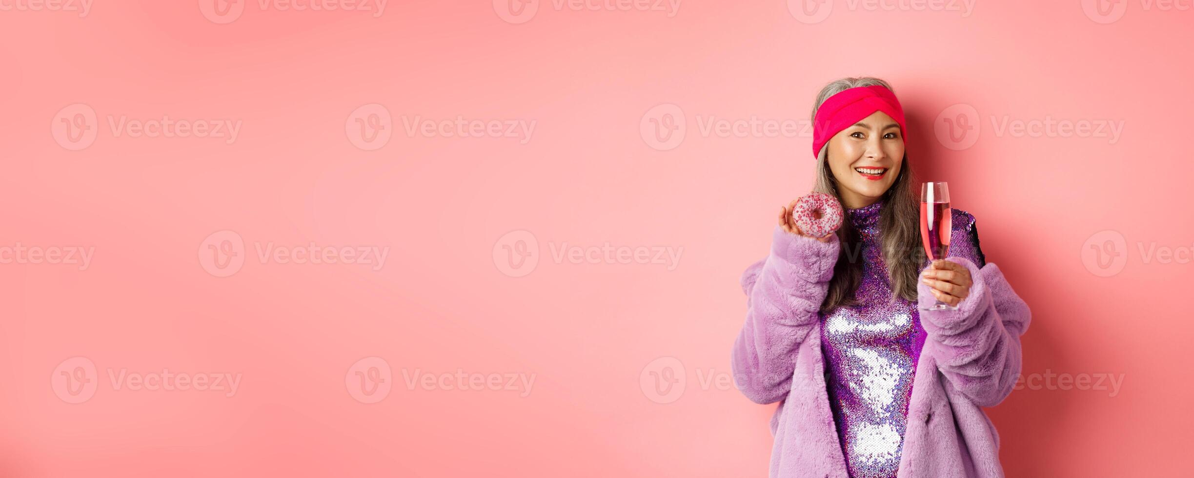 Elegant and stylish asian woman in purple faux fur coat eating donuts and drinking champagne, having fun at party, standing over pink background photo