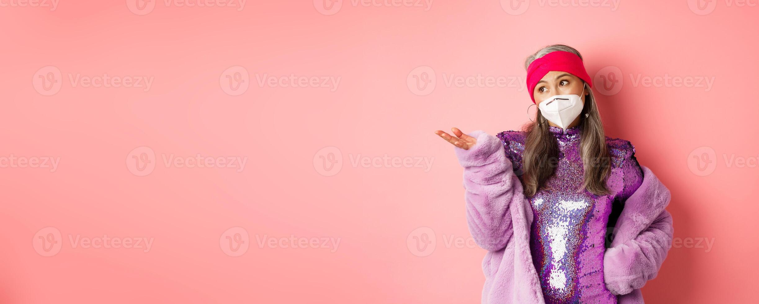 Covid-19, pandemic and fashion concept. Stylish asian senior woman in respirator and party dress looking skeptical, pointing hand left, standing over pink background photo
