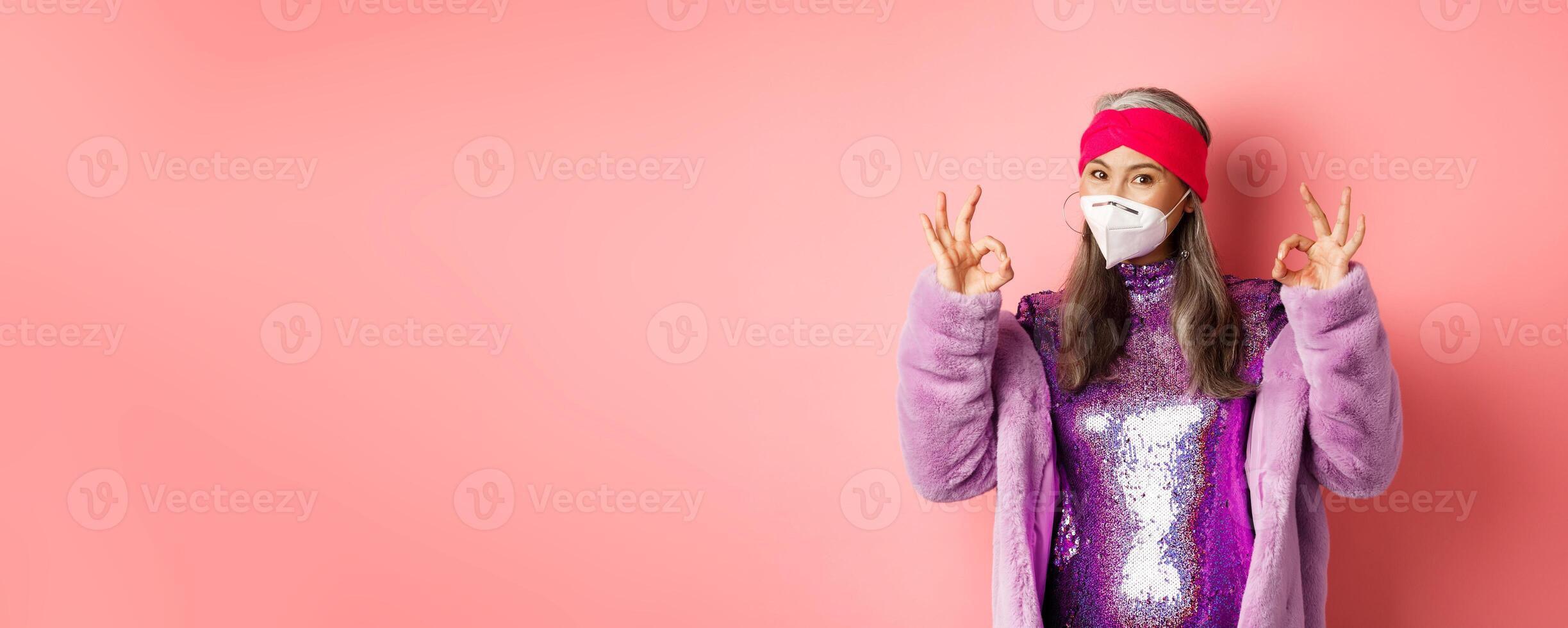 Covid-19, pandemic and fashion concept. Cool asian grandmother wearing stylish disco dress and respirator, showing okay signs, asking to wear face masks and social distancing, pink background photo