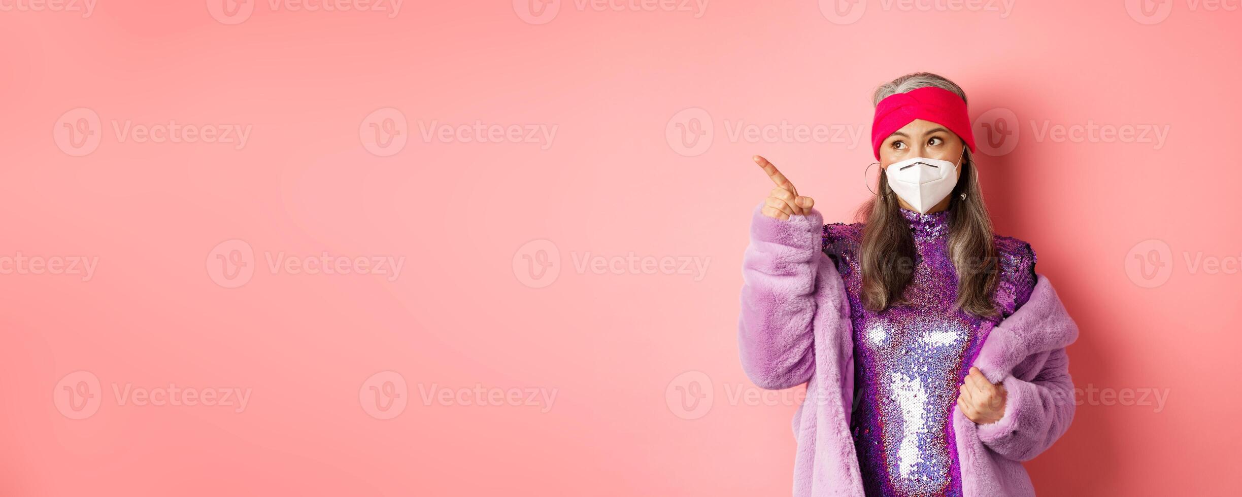 Covid and fashion concept. Stylish asian senior woman checking out special deal, wearing disco dress and face mask, pointing finger left at copy space, pink background photo