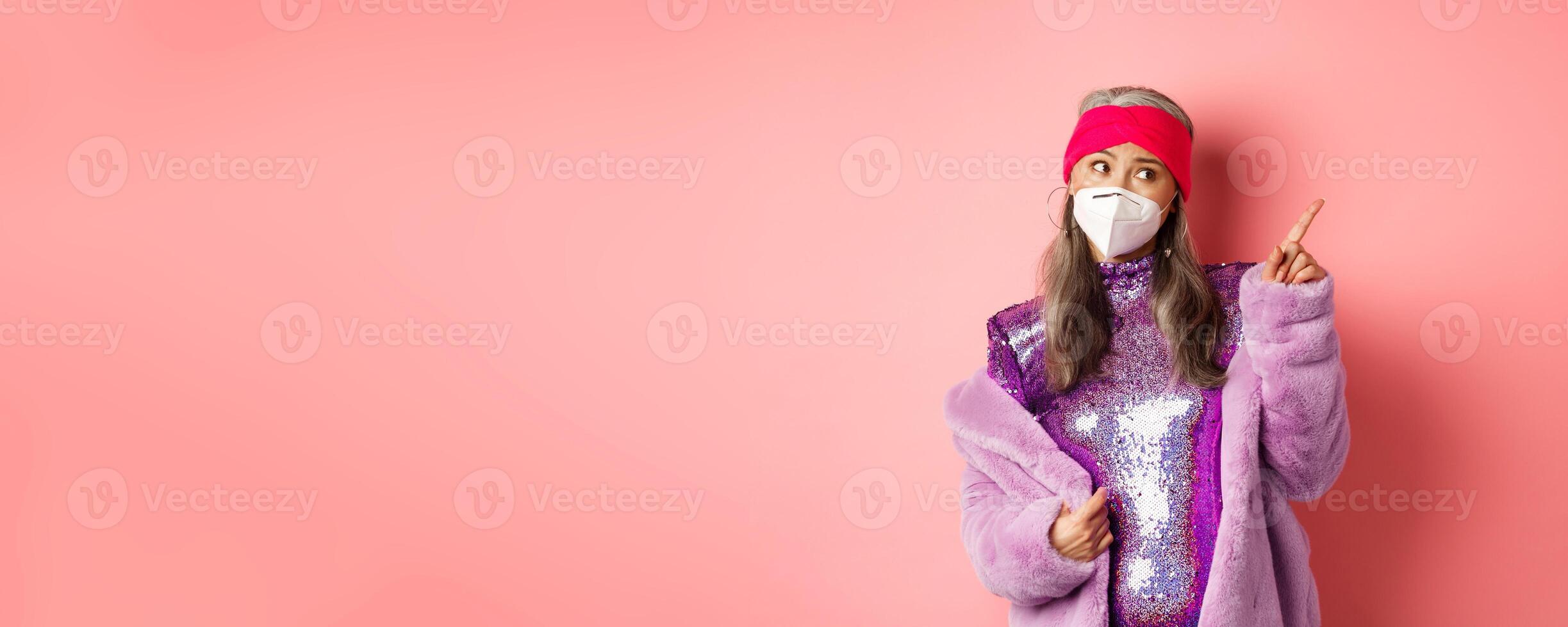 Coronavirus and shopping concept. Sassy elderly asian woman in respirator and glittering shiny dress, pointing at upper right corner and look doubtful, pink background photo