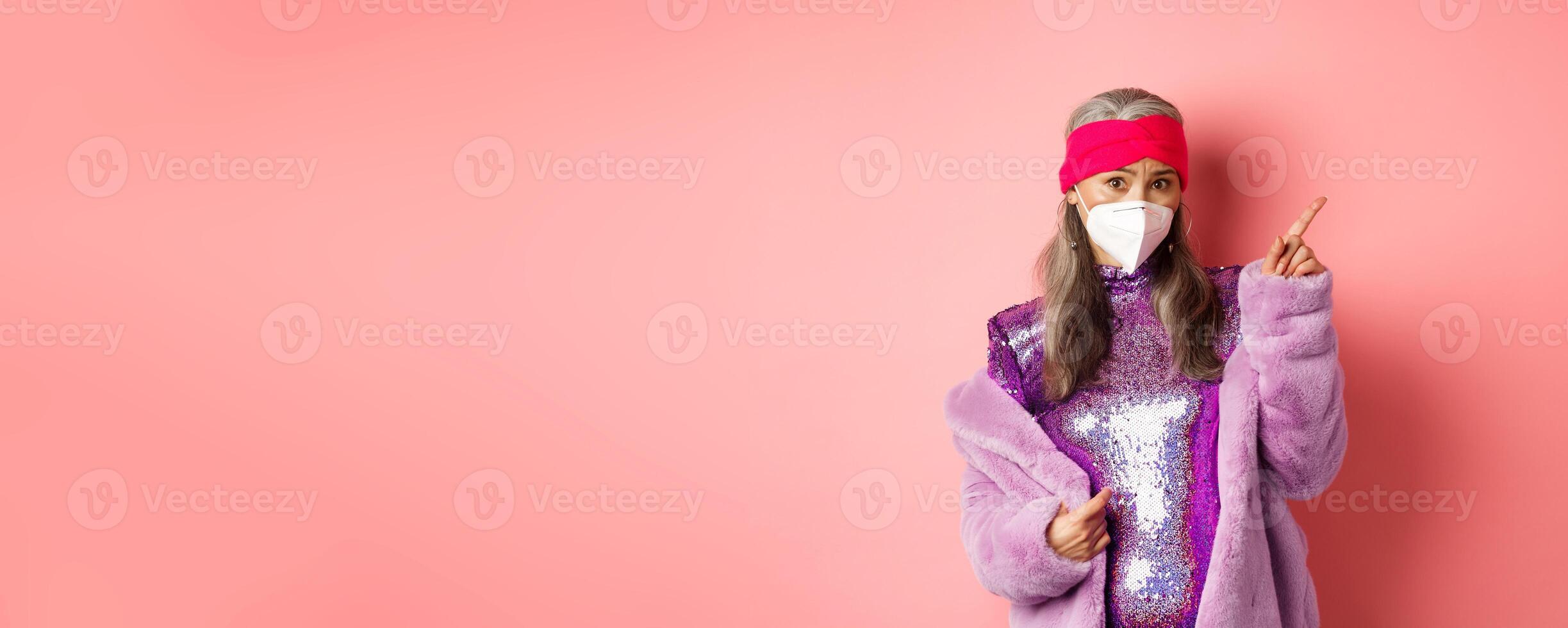 Coronavirus and shopping concept. Worried asian senior woman in stylish outfit and respirator pointing upper right corner, looking unsure at camera, asking advice, pink background photo
