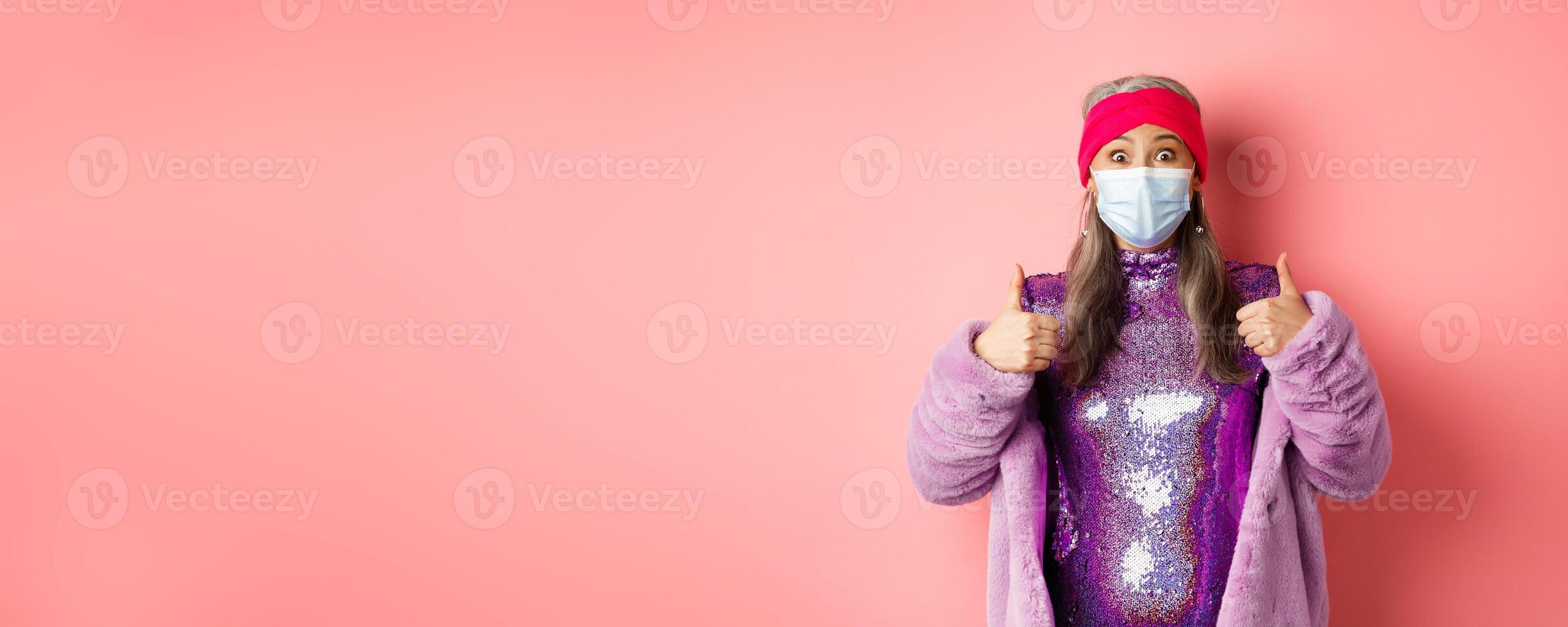 Covid-19, social distancing and fashion concept. Impressed asian senior woman in party outfit and respirator showing thumbs-up, check out awesome special deal, pink background photo