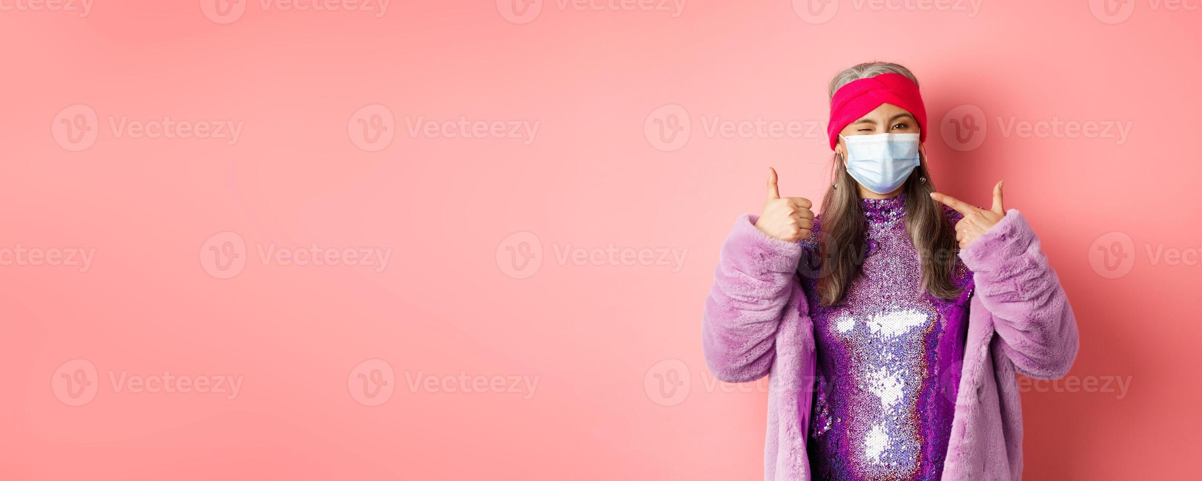 Covid-19, social distancing and fashion concept. Smiling asian senior woman in fashionable clothes and respirator, pointing at face mask and show thumb-up, pink background photo