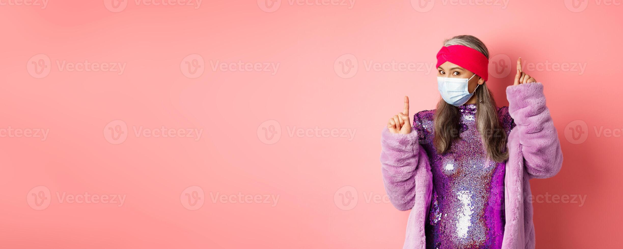 Covid-19, social distancing and fashion concept. Cheerful asian senior woman likes to party, dancing in respirator and shiny purple dress, standing over pink background photo