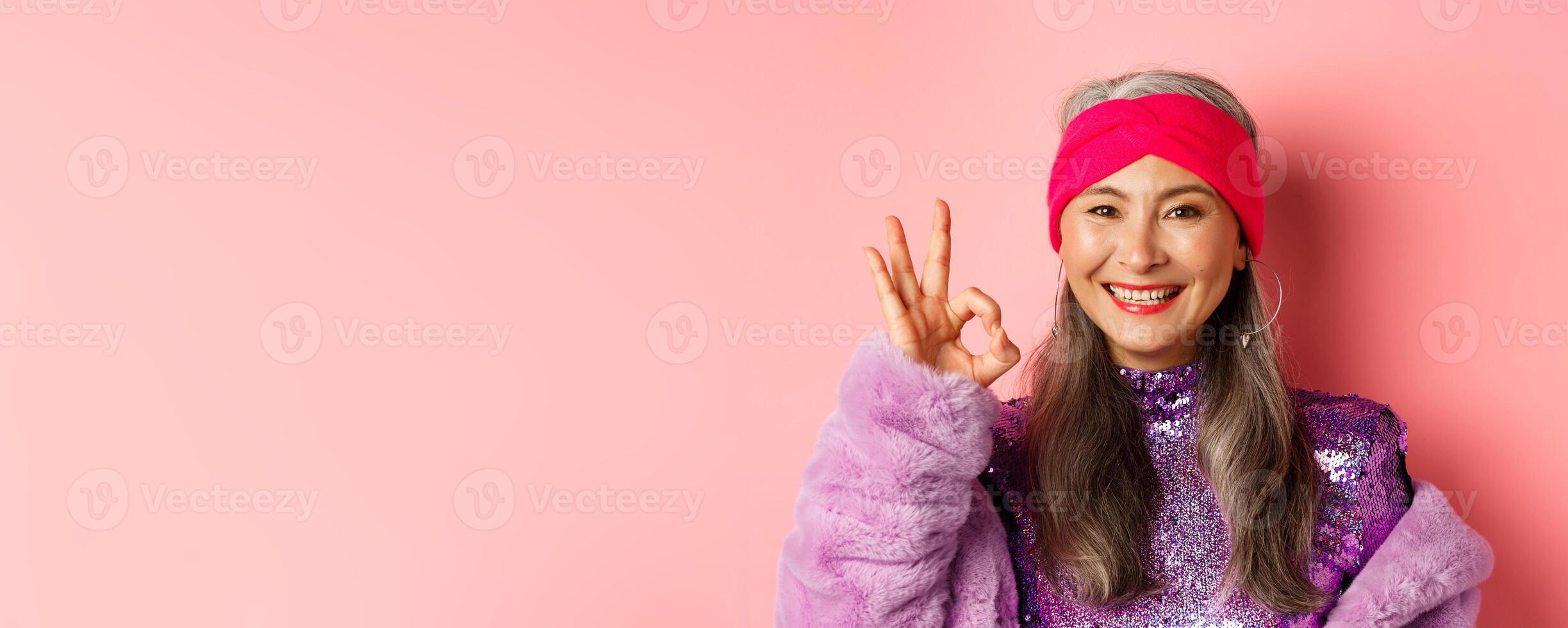 Fashion. Close-up of asian old lady in stylish disco outfit showing OK sign, approve and like something, smiling pleased, pink background photo