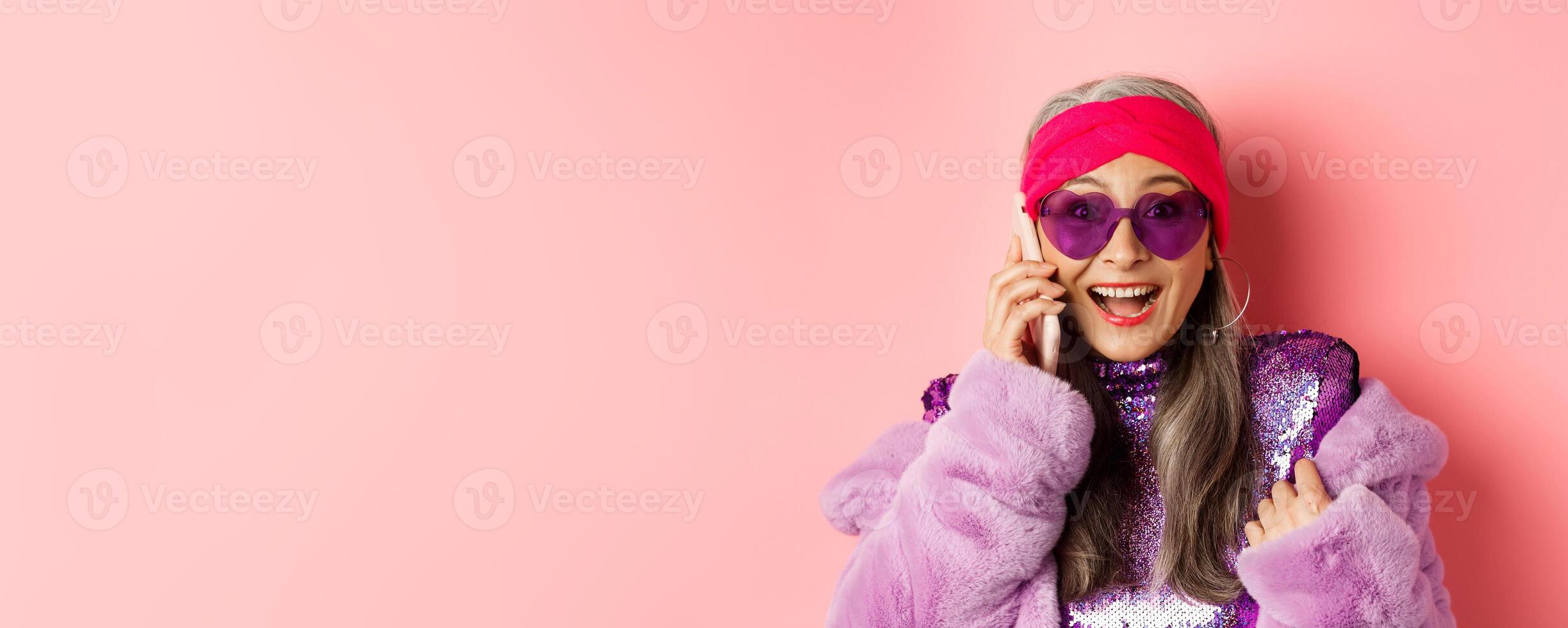 Fashion. Close-up of stylish asian senior woman hear amazing news on mobile phone, smiling happy at camera, wearing trendy sunglasses, pink background photo