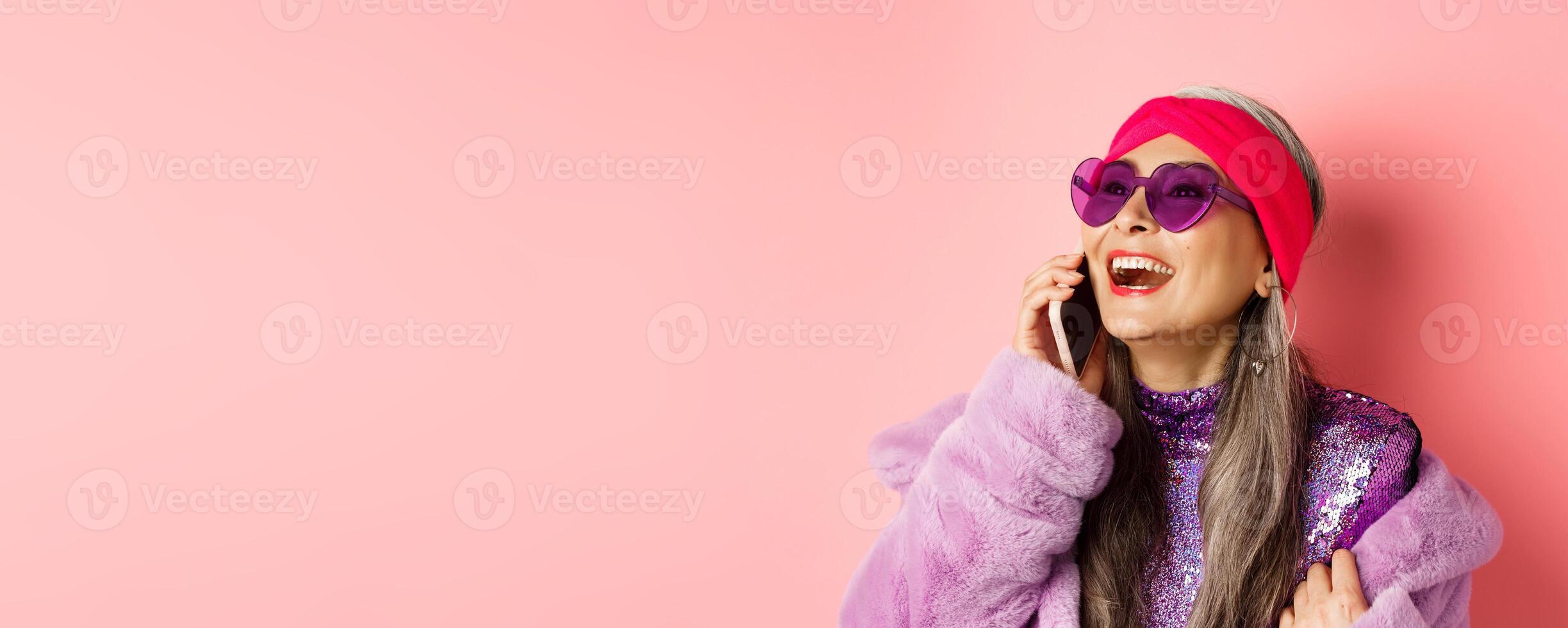 Fashion. Beautiful asian senior woman in trendy sunglasses and headband laughing, talking on mobile phone and looking aside, pink background photo