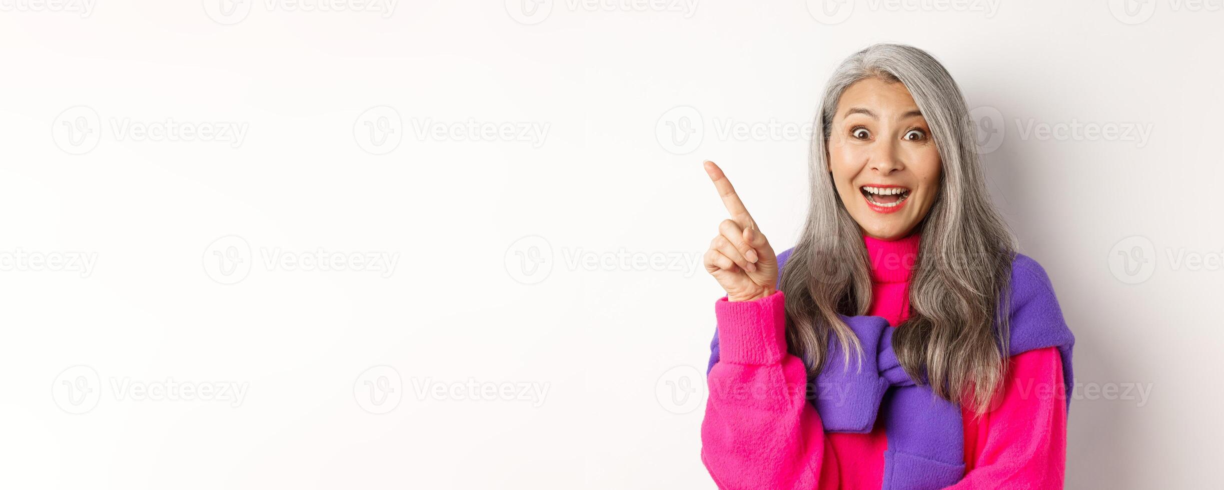 Close up of beautiful asian female model pointing finger at upper left corner, smiling and looking happy at camera, showing promo deal, white background photo