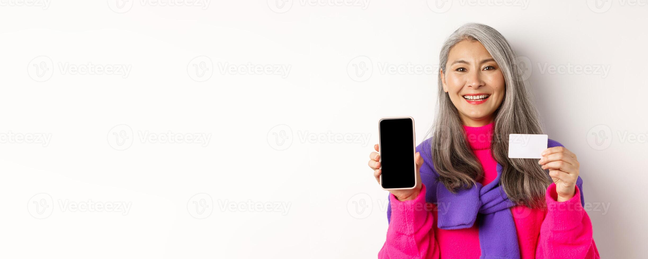 Online shopping. Closeup of smiling asian senior woman showing blank mobile screen and plastic credit card, looking happy, standing over white background photo