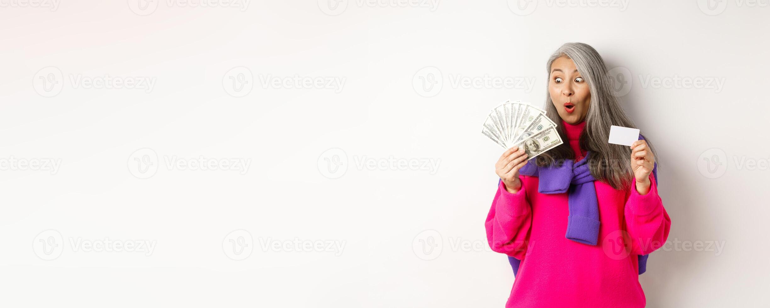 Shopping concept. Lucky asian senior woman looking amazed at money and showing plastic credit card, standing over white background photo