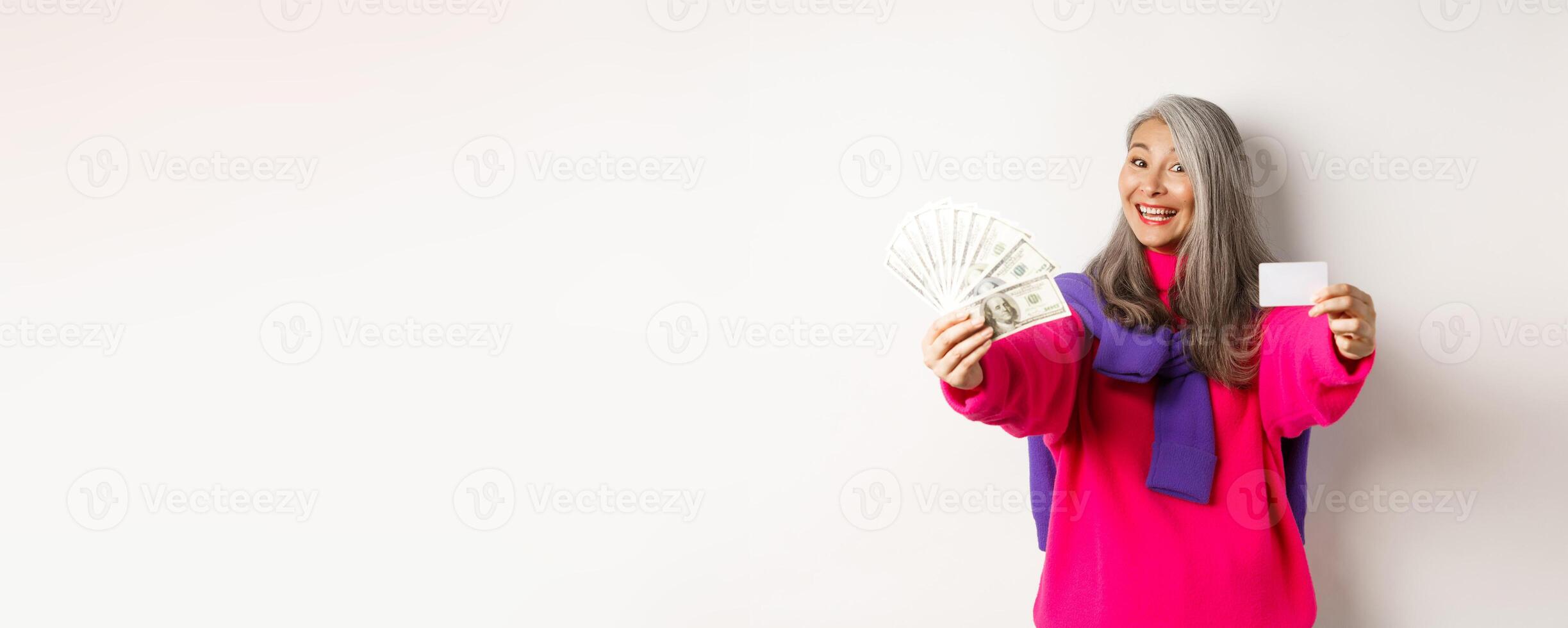 Shopping concept. Cheerful asian senior woman showing cash and plastic credit card, smiling at camera, standing over white background photo