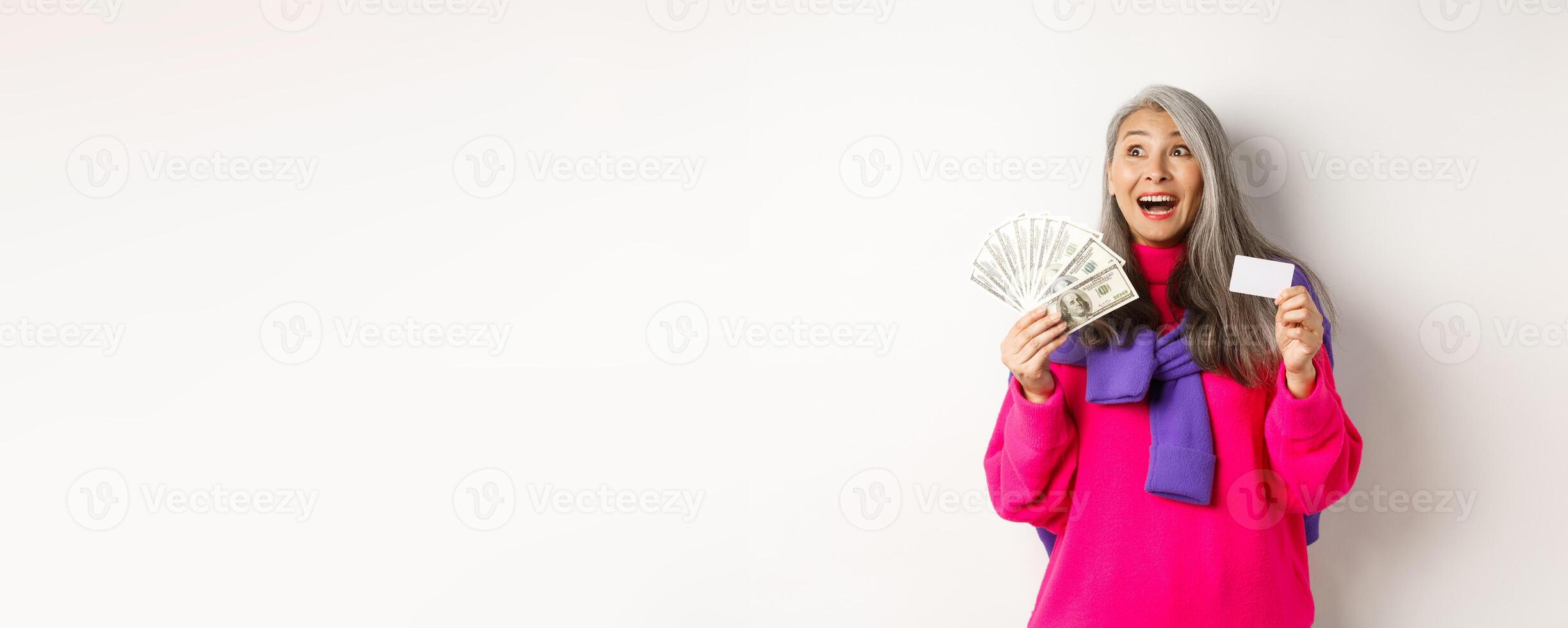 Shopping concept. Happy asian old woman with grey hair, looking fascinated at upper left corner, showing plastic credit card and money dollars, white background photo