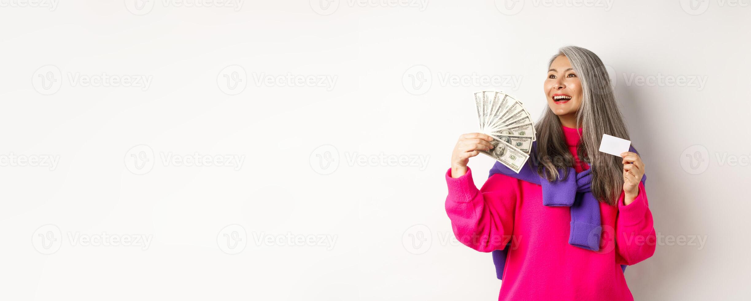 Shopping concept. Stylish asian senior woman showing money dollars and plastic credit card, looking dreamy aside, thinking about buying, white background photo