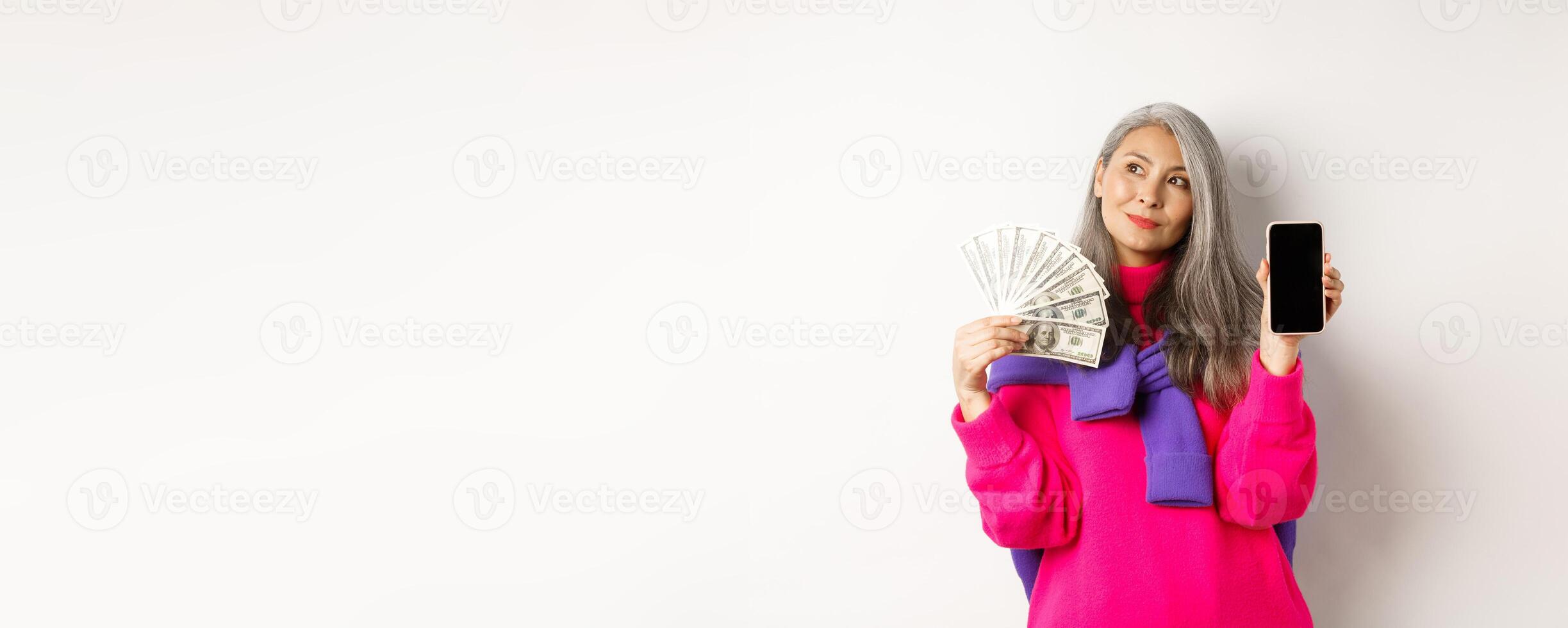 Fashionable asian senior woman showing money dollars and blank smartphone screen, demonstrate online shop, standing over white background photo