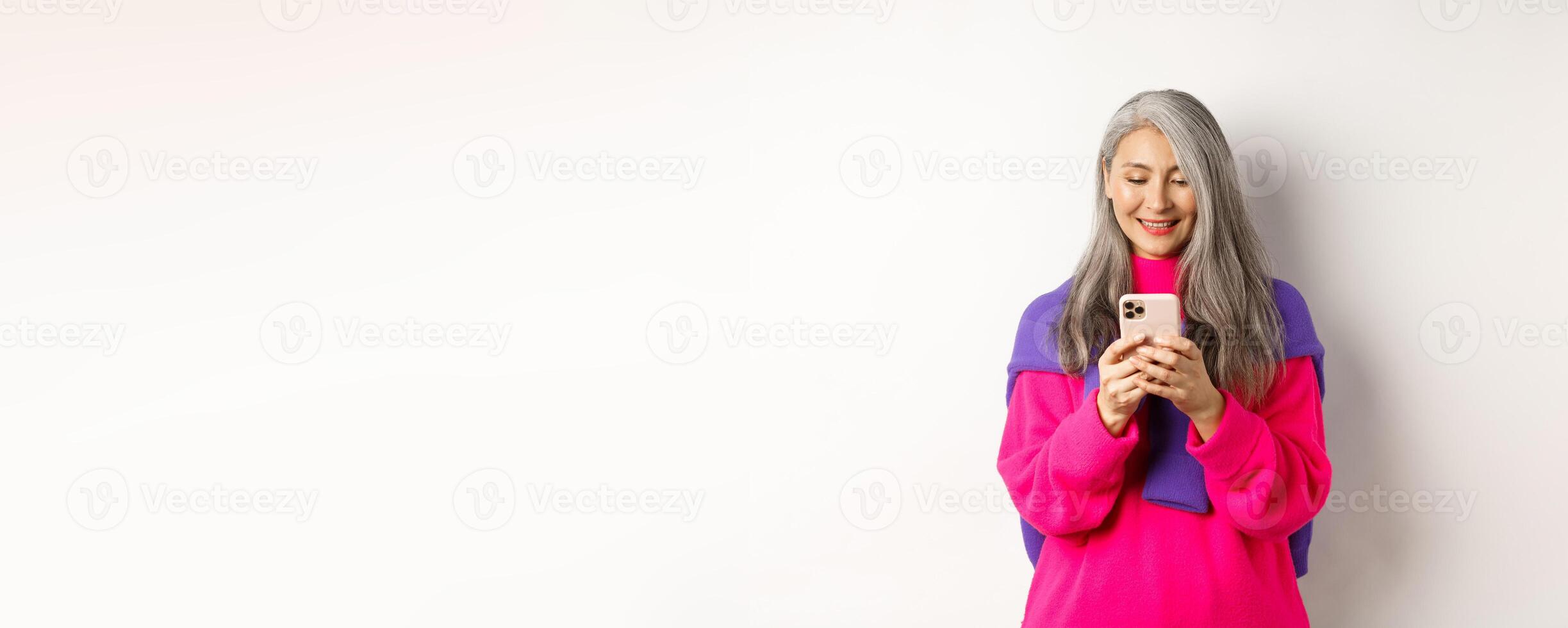 Beautiful asian senior woman in pink stylish sweater using smartphone. Old lady reading on mobile phone and smiling, standing over white background photo
