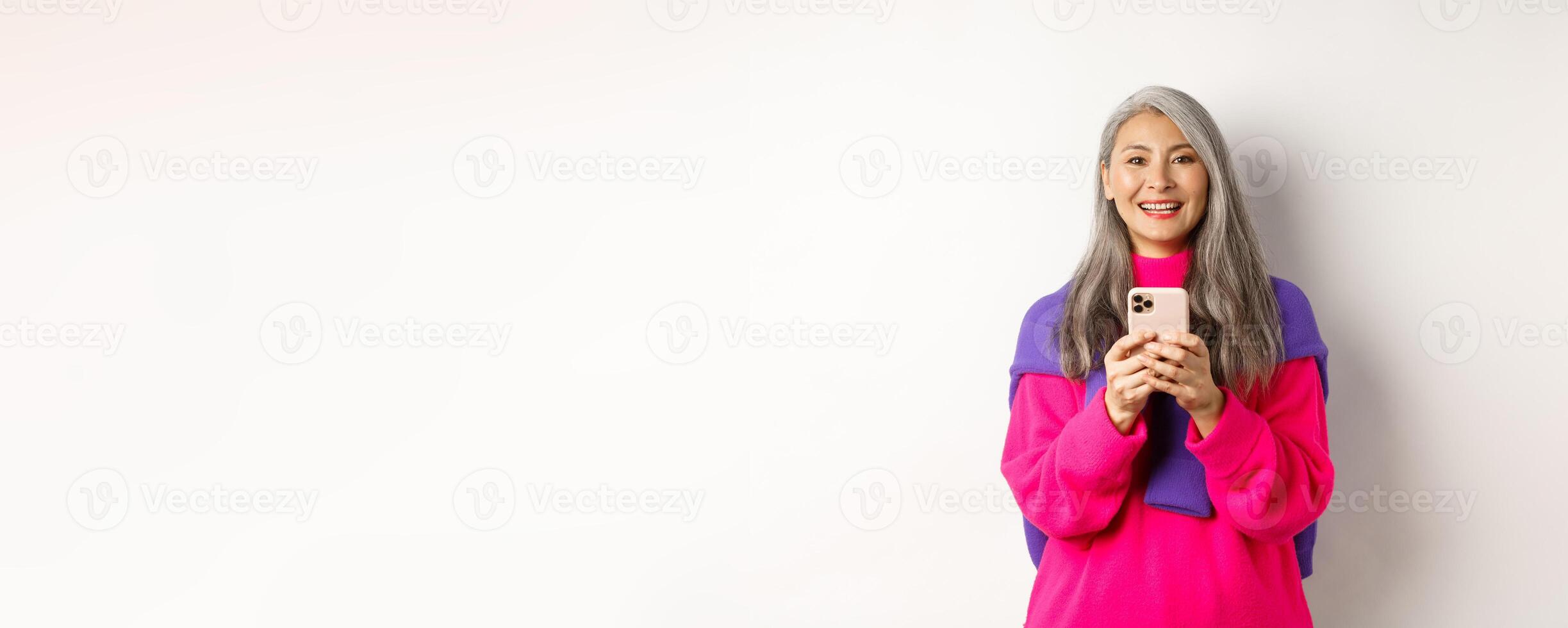 Happy asian woman smiling at camera after reading promotion on smartphone, standing with mobile phone over white background photo