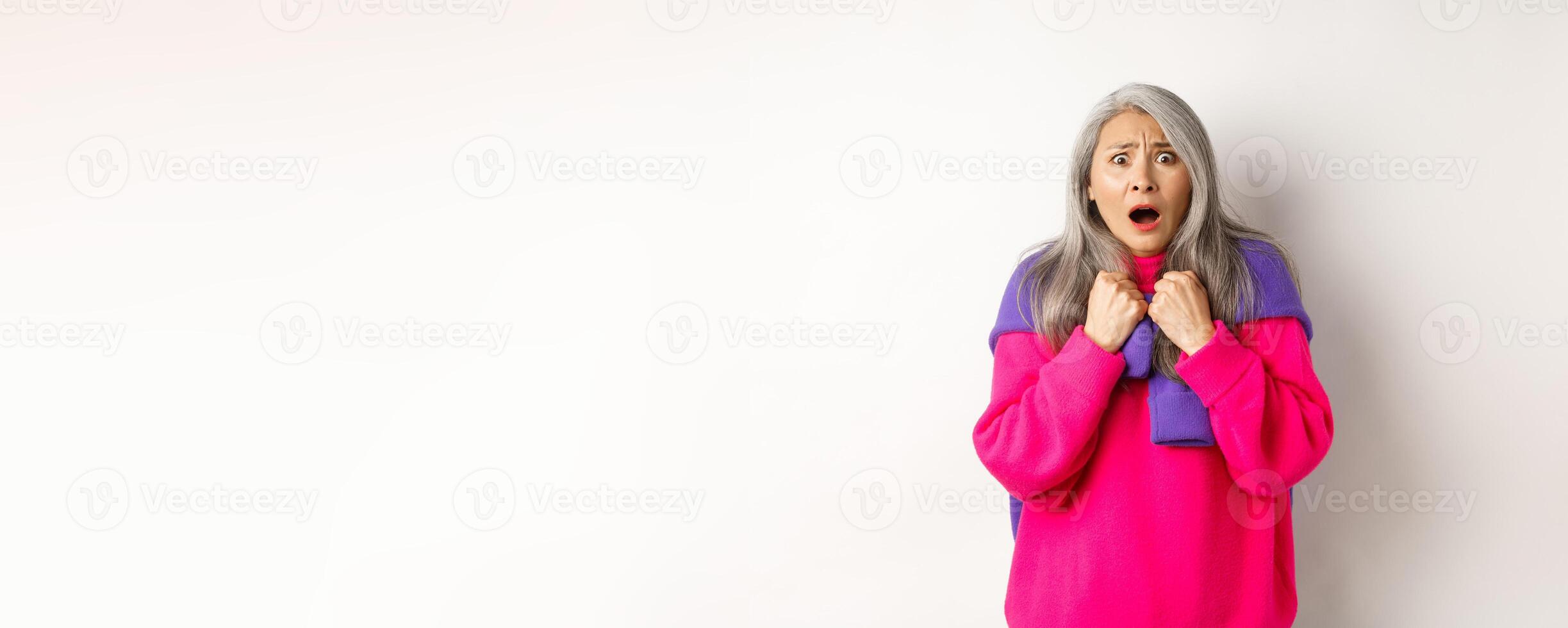 Scared and shocked asian grandmother gasping and staring frustrated at camera, standing against white background photo