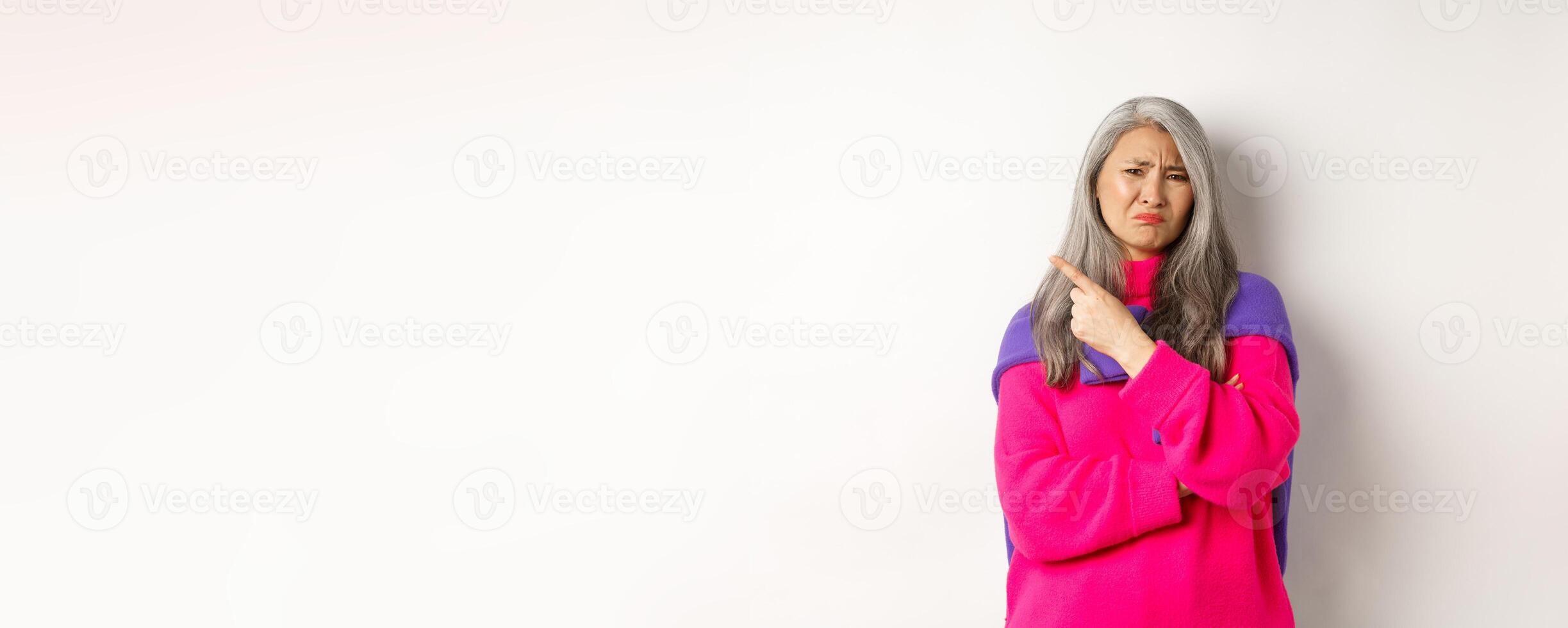 Disappointed asian grandmother grimacing disgusted, pointing finger left and complaining on something bad, standing over white background photo