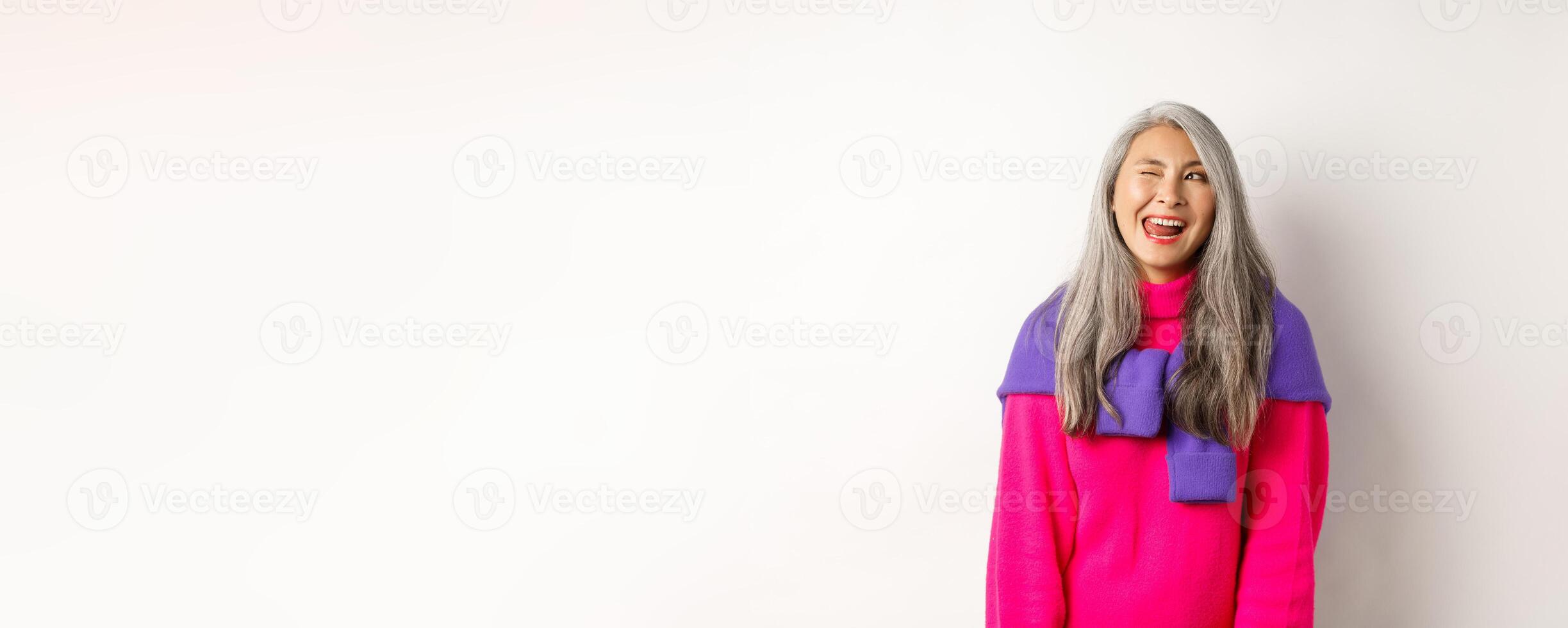 Funny and happy asian senior woman having fun, showing tongue and winking cheeky, looking left at logo, standing over white background photo