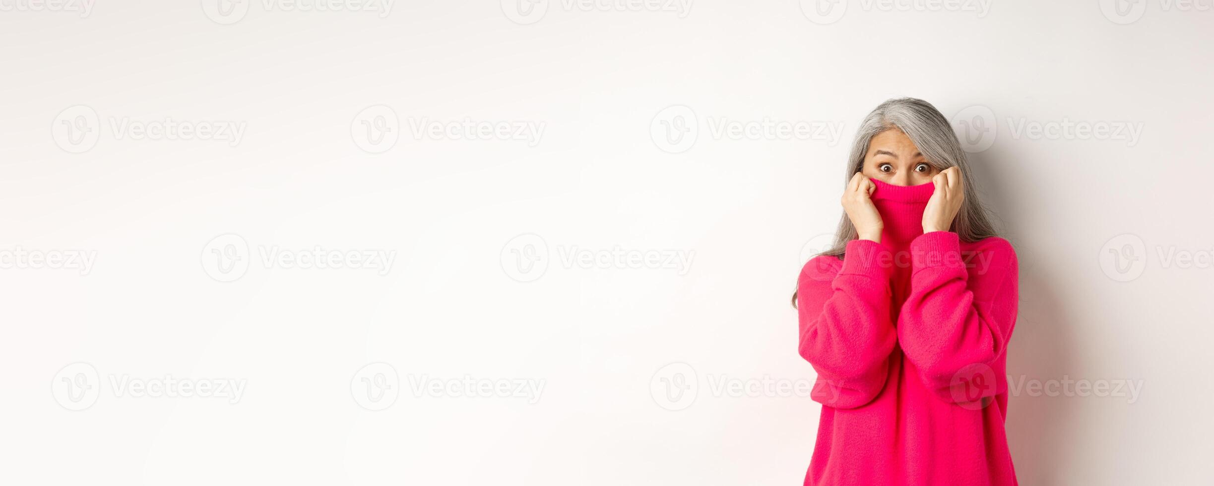 Portrait of funny asian grandmother hiding face in sweater collar, peeking at camera silly, standing over white background photo