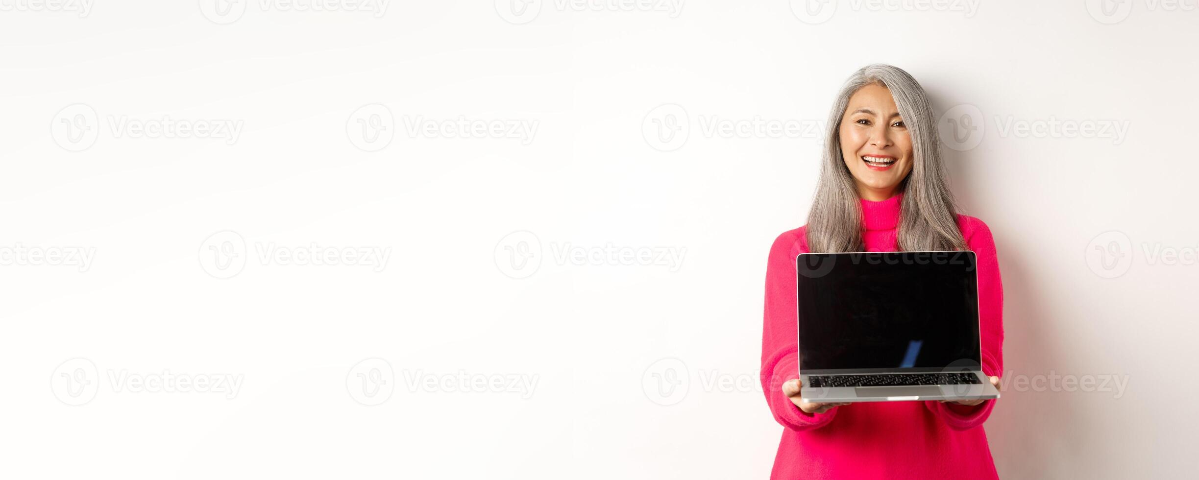 E-commerce concept. Beautiful asian senior businesswoman showing blank laptop screen and smiling, standing in pink sweater over white background photo
