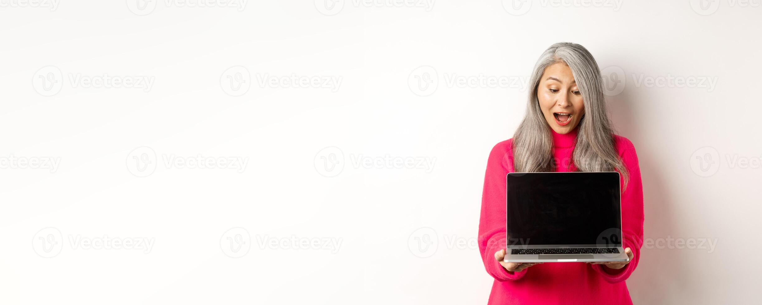 E-commerce concept. Amazed asian grandmother with grey hair, checking out promo online, showing laptop black screen, standing over white background photo