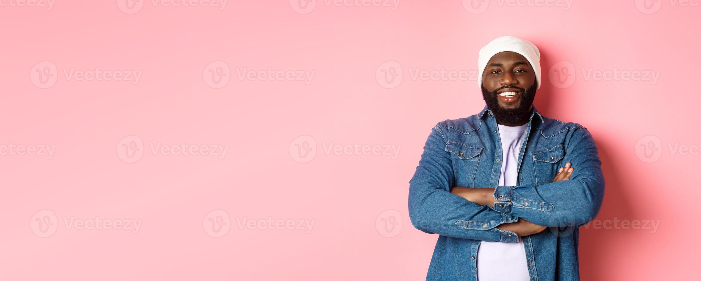 Handsome hip hop style Black man in beanie and denim shirt, smiling confident, cross arms on chest and staring at camera on pink background photo