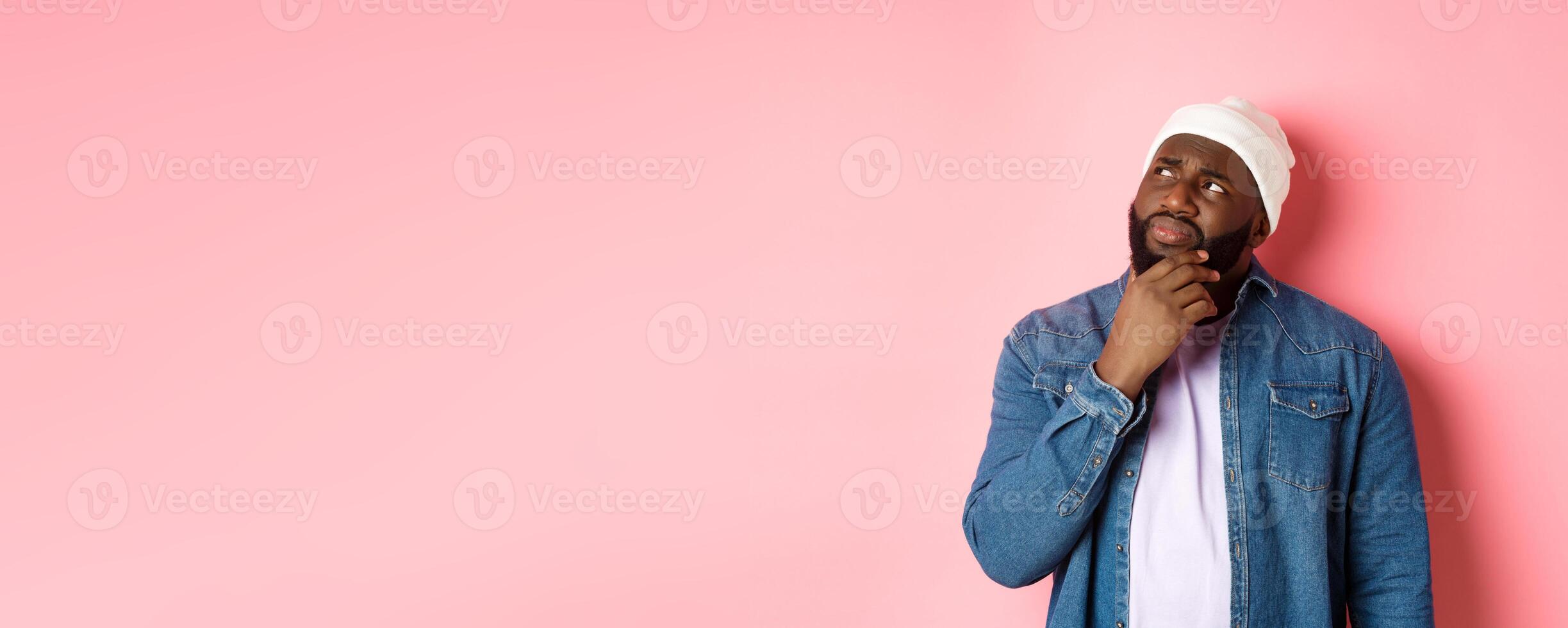 Image of puzzled and concerned black man, staring at upper left corner and thinking, making decision with doubts, standing over pink background photo