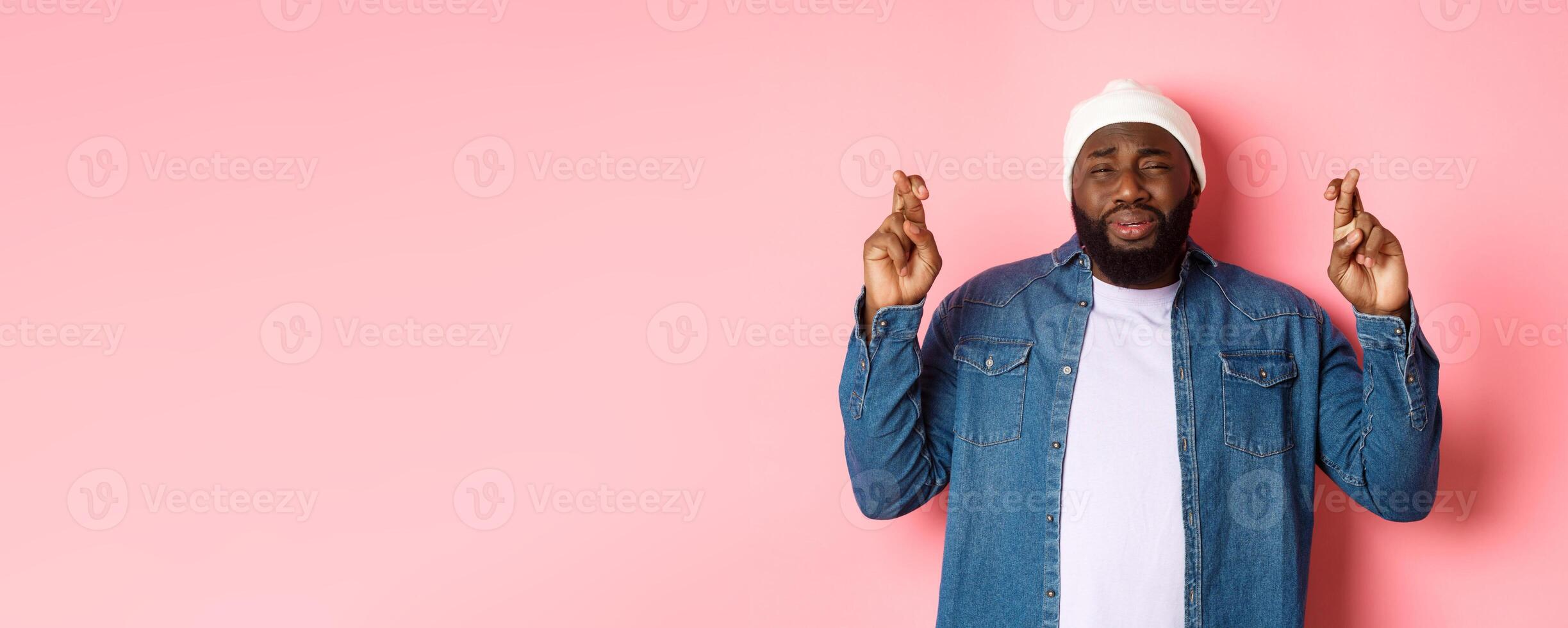 Image of young african-american man whining, pleading god, cross fingers and crying, standing over pink background photo