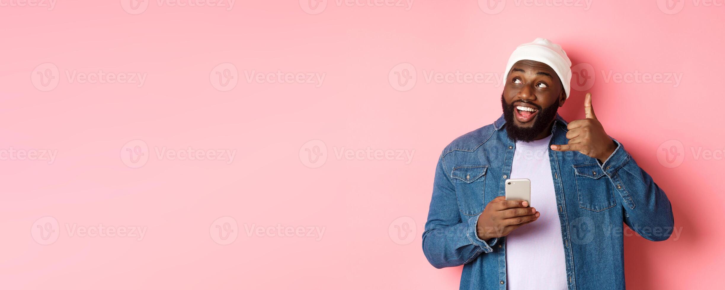Happy Black man showing call me sign, making phone gesture and smiling, holding smartphone, standing in beanie and denim shirt over pink background photo