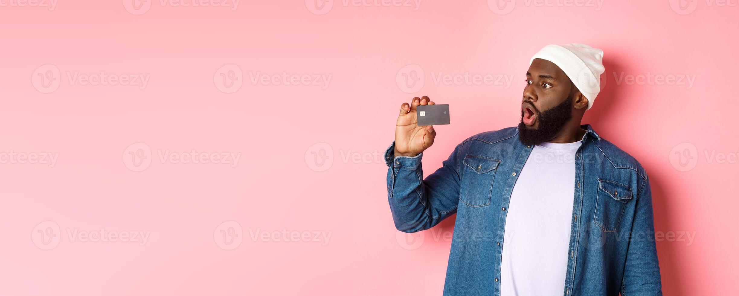 Shopping concept. Image of bearded african-american man staring shocked at credit card, standing against pink background photo