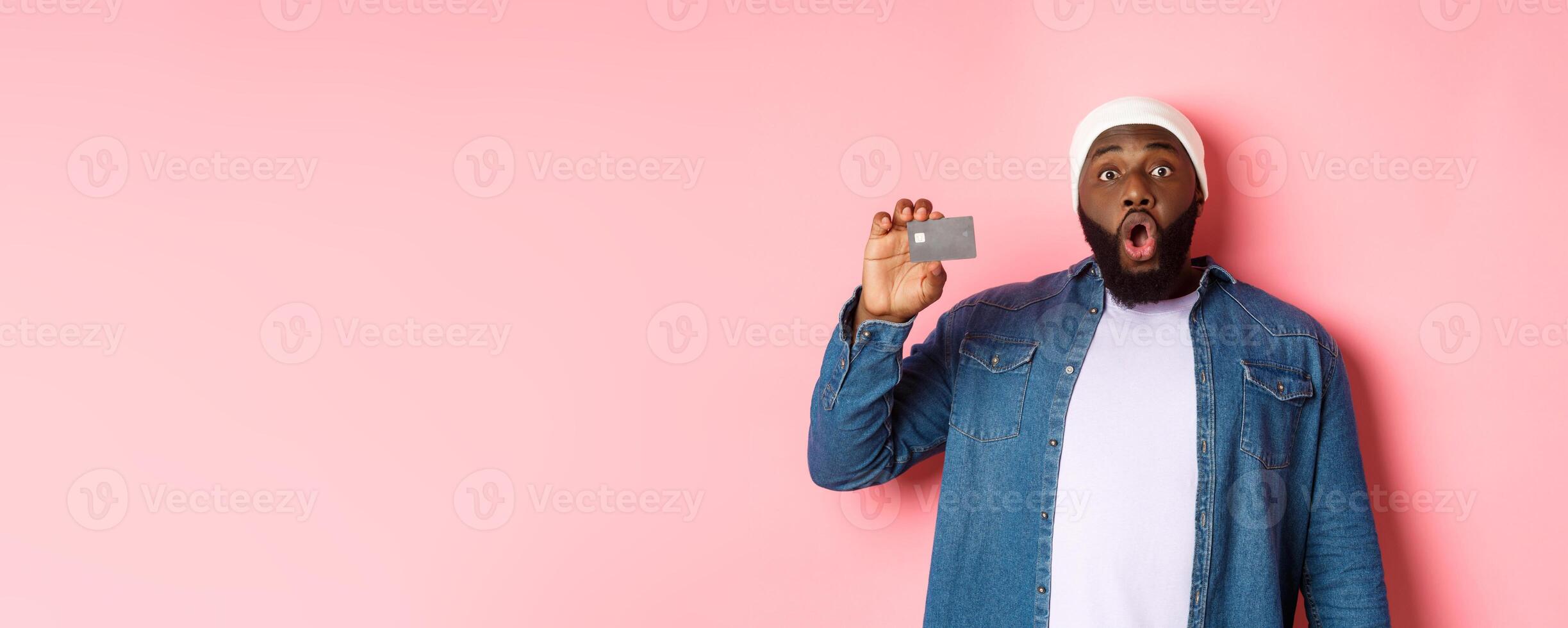 Shopping concept. Amazed Black guy showing credit card, staring at camera impressed, recommending bank, standing over pink background photo
