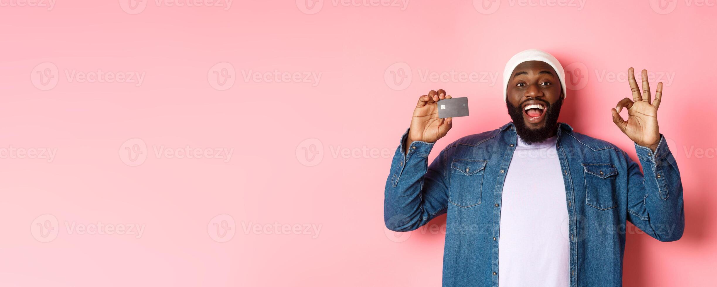 Shopping concept. Handsome african-american man recommending bank, showing credit card and okay sign, smiling satisfied, standing over pink background photo