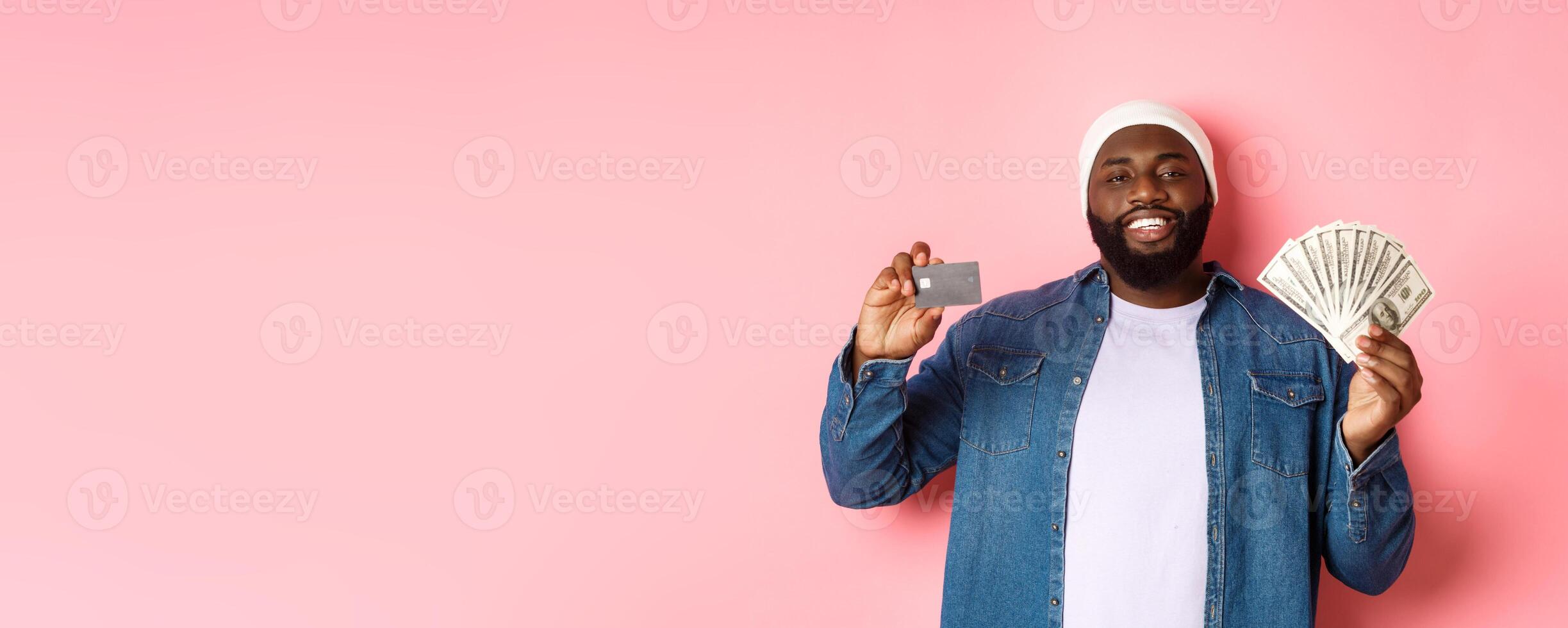 Shopping concept. Handsome young Black guy showing credit card of bank and money, smiling satisfied, standing over pink background photo