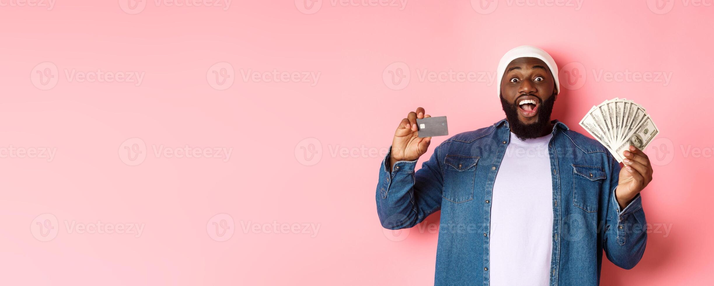 Shopping concept. Excited african-american man showing credit card and dollars, got deposit or money loan, standing over pink background photo