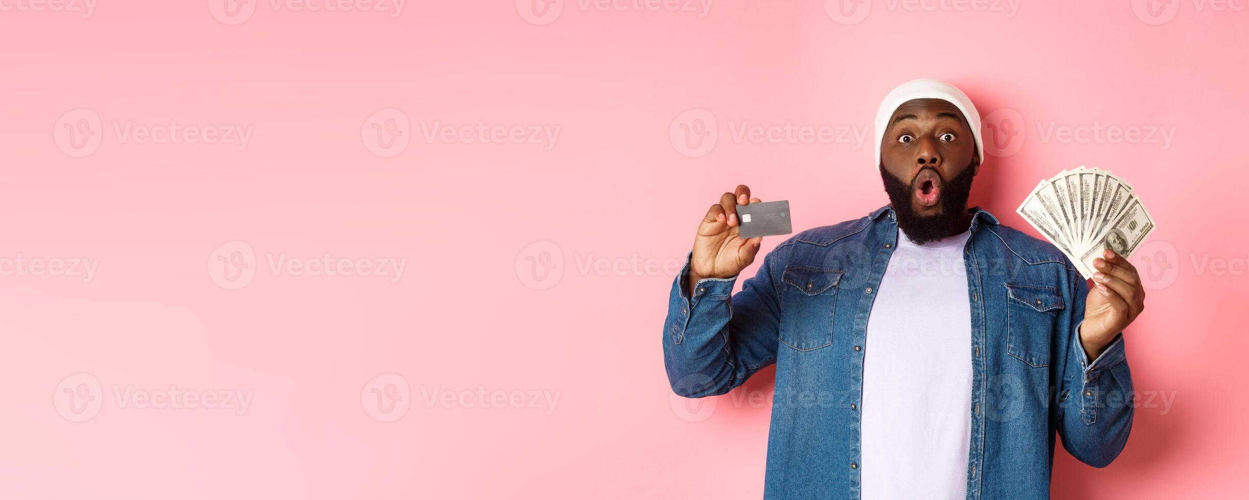 Shopping concept. Excited african-american man showing credit card and dollars, got deposit or money loan, standing over pink background photo