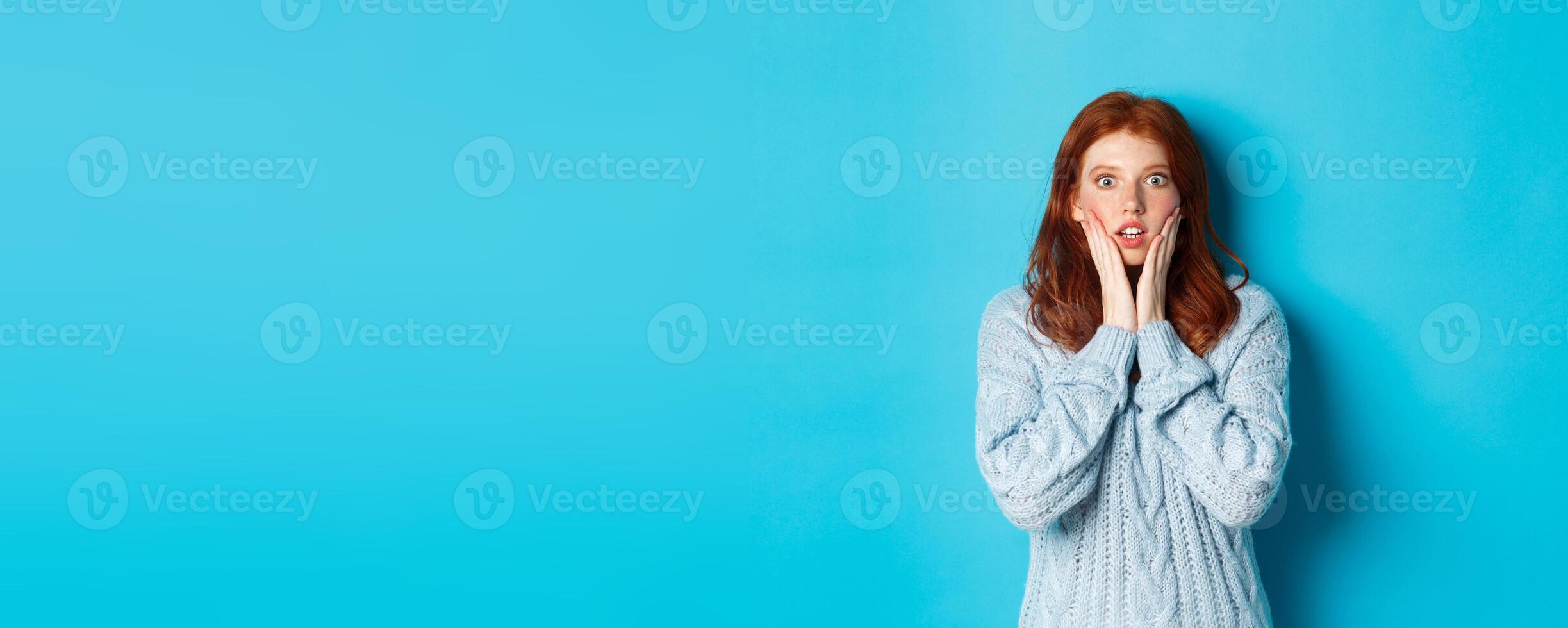 Shocked redhead girl staring at camera speechless, express disbelief and amazement, standing in sweater against blue background photo