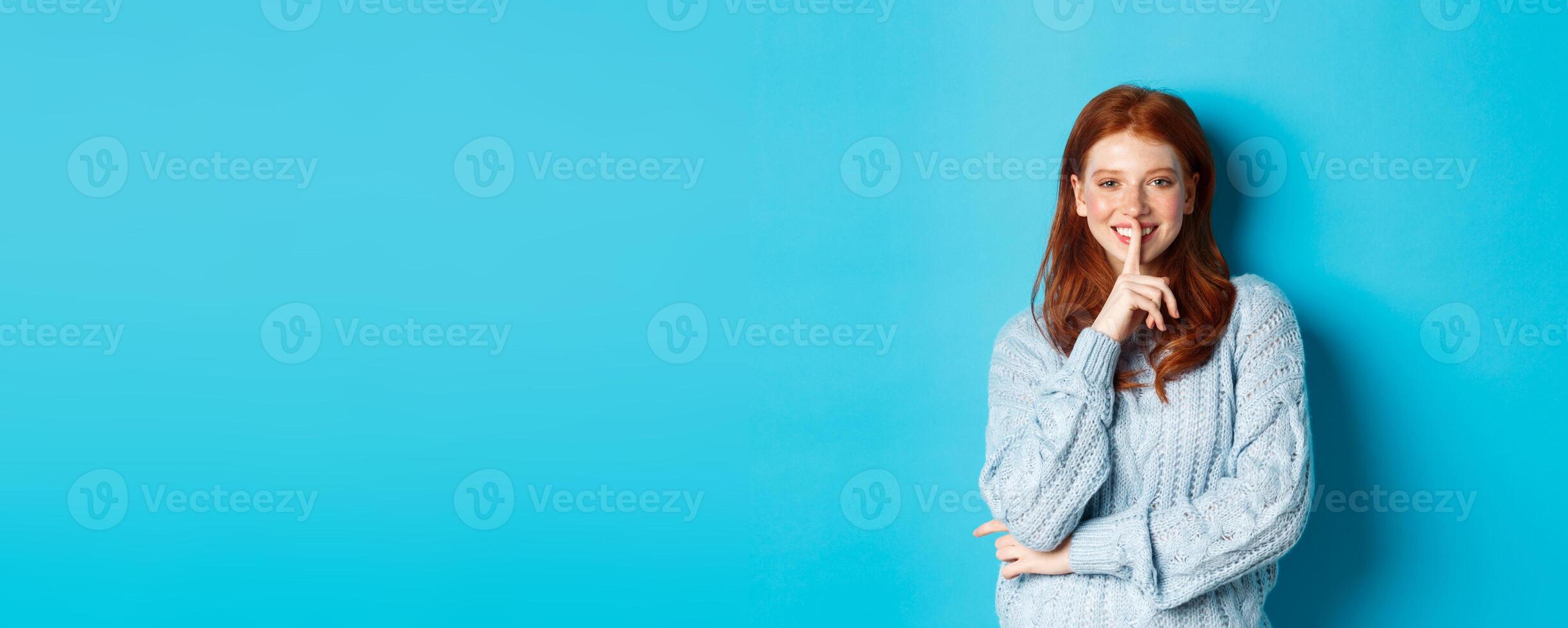 Pretty redhead teenager hushing and smiling, telling a secret, standing in sweater against blue background photo