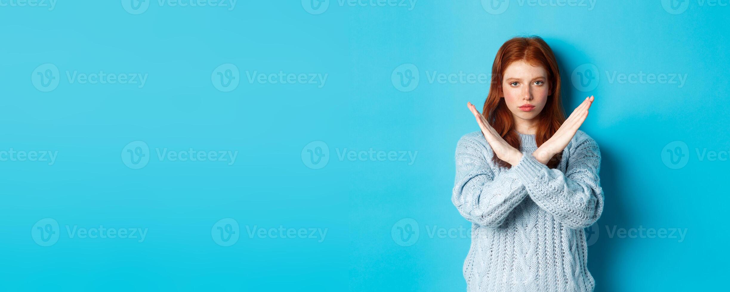 Serious redhead girl looking confident, showing cross gesture to stop and forbid action, standing over blue background photo