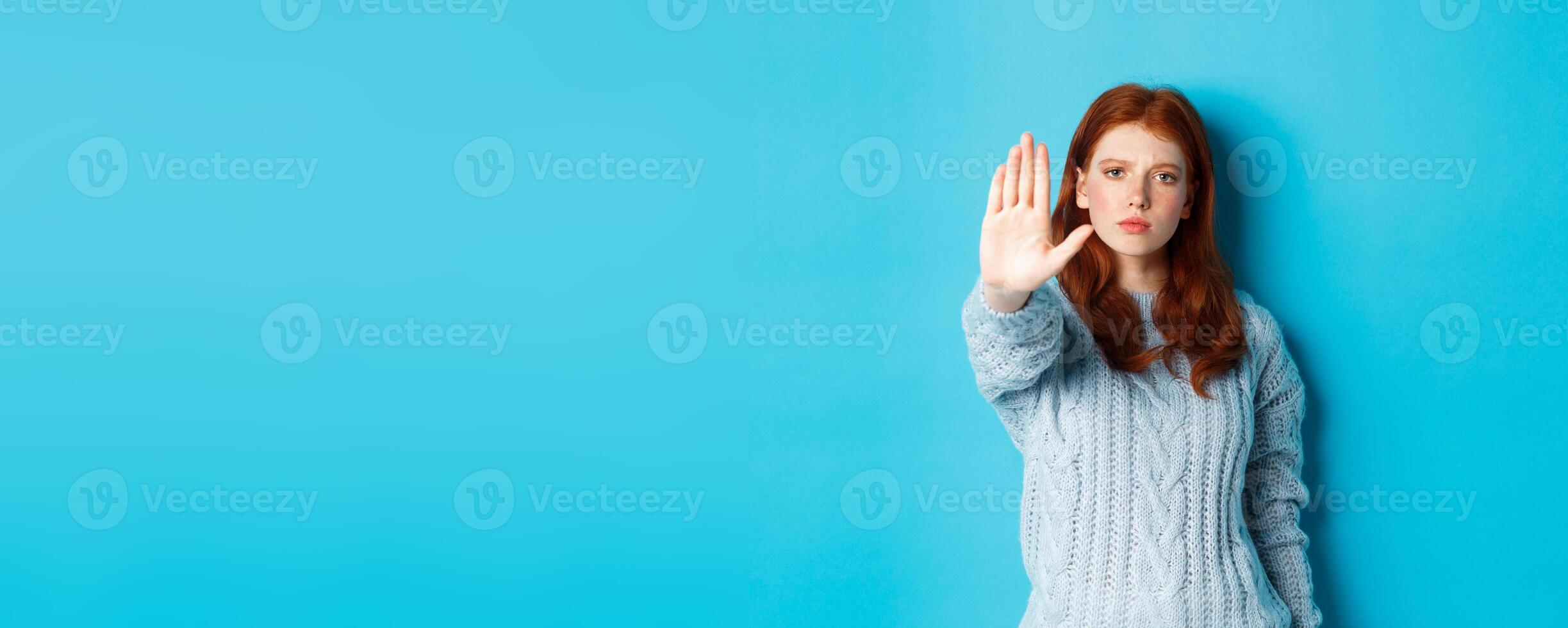 Serious and confident redhead girl telling to stop, saying no, showing extended palm to prohibit action, standing over blue background photo