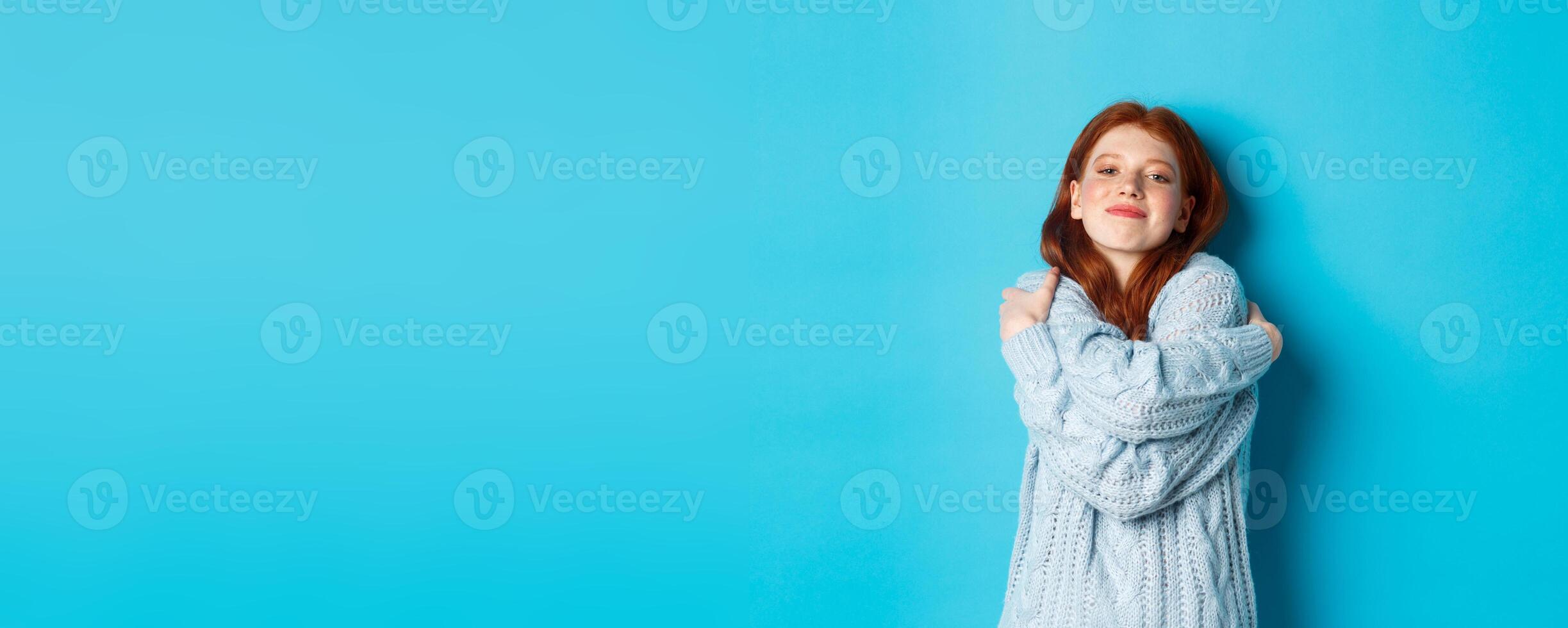 Happy cute redhead girl hugging herself, wearing comfortable and warm sweater, smiling at camera, standing over blue background photo