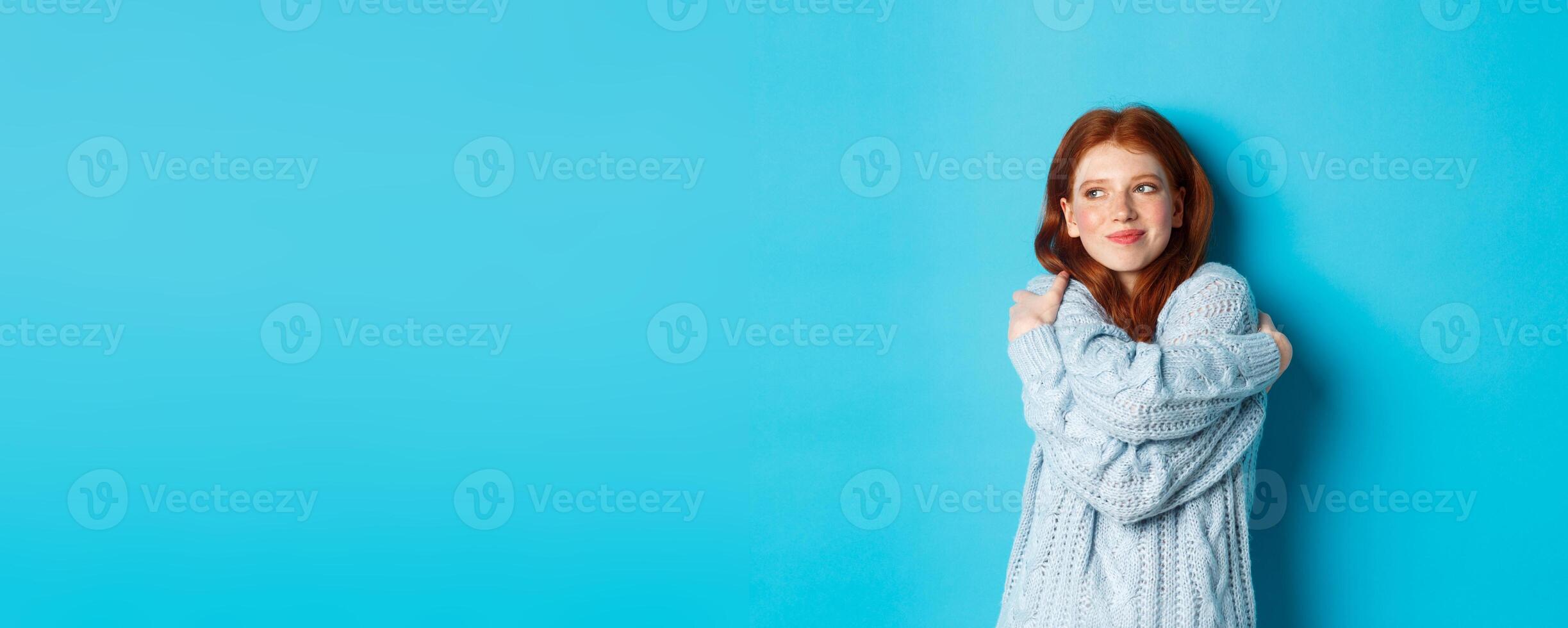 Happy cute redhead girl hugging herself, wearing comfortable and warm sweater, smiling at camera, standing over blue background photo