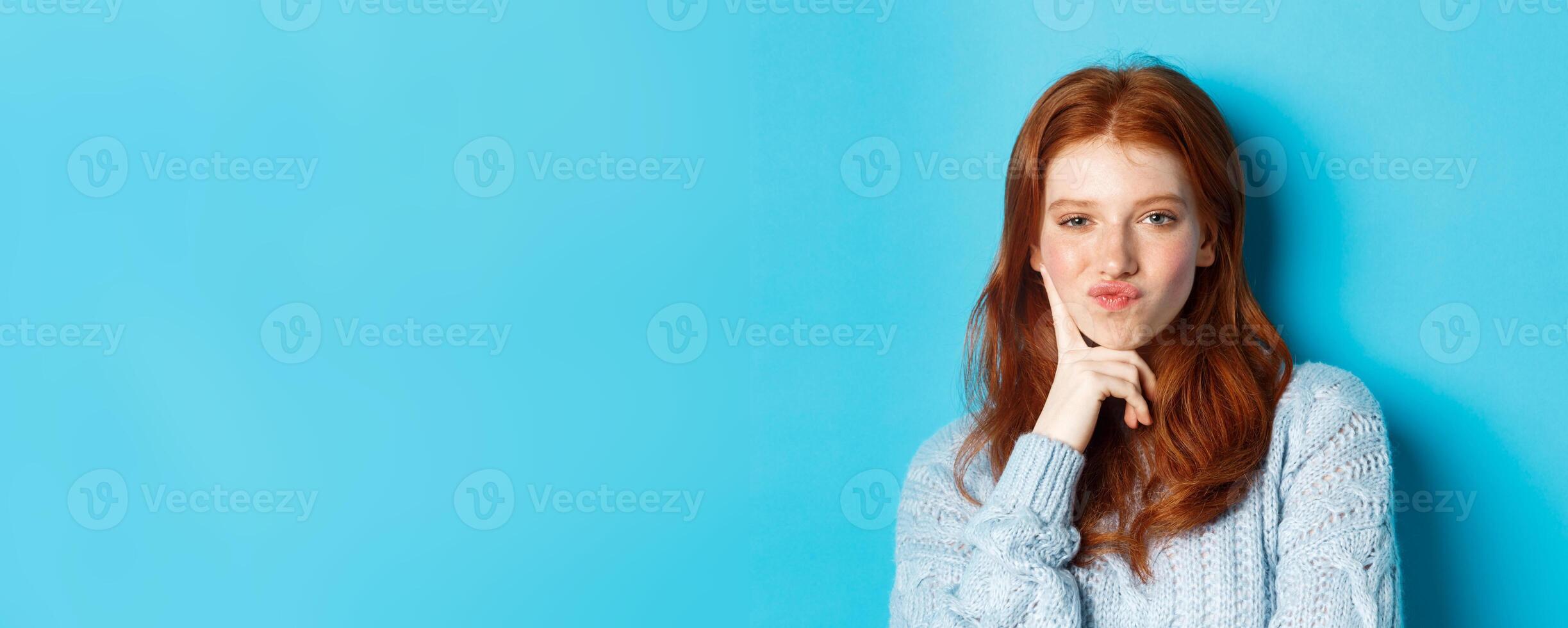 Close-up of beautiful redhead girl thinking, pucker lips and staring thoughtfula t camera, making choice, standing over blue background photo