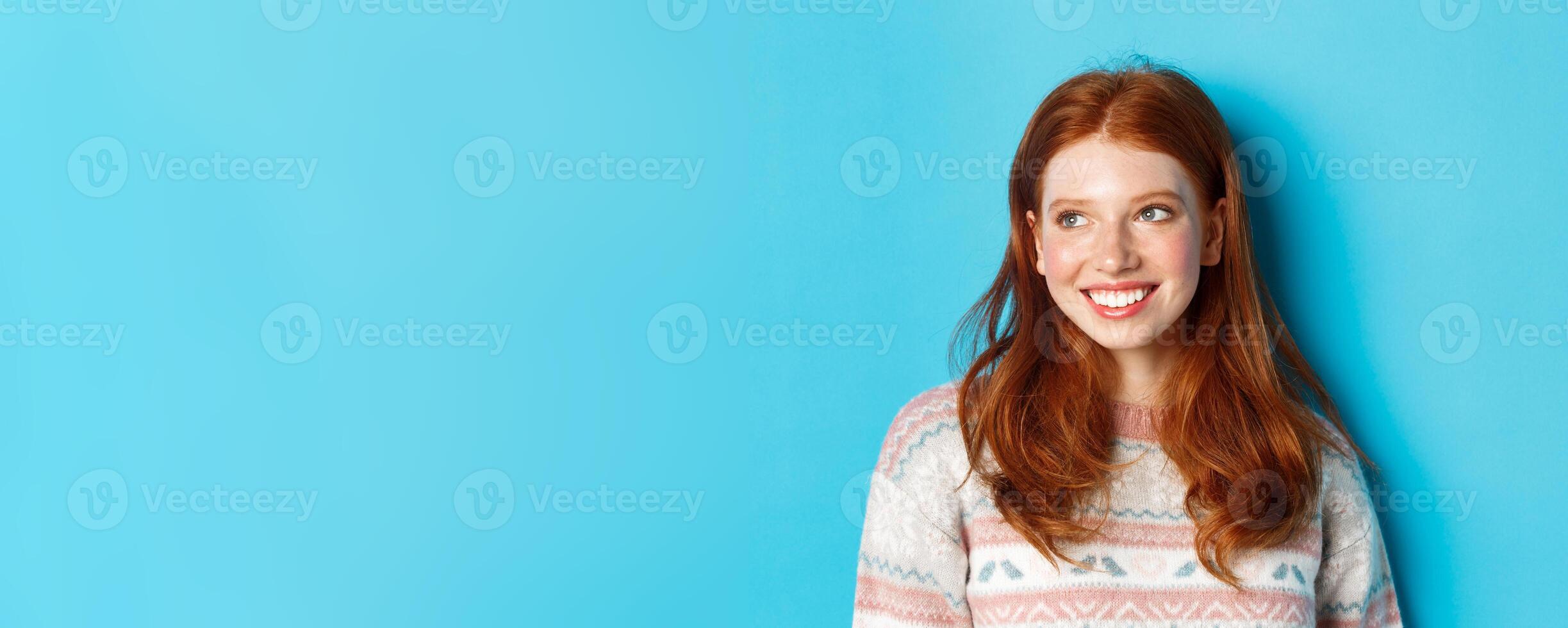 Close-up of beautiful redhead female model in winter sweater, smiling and looking left at copy space, standing over blue background photo