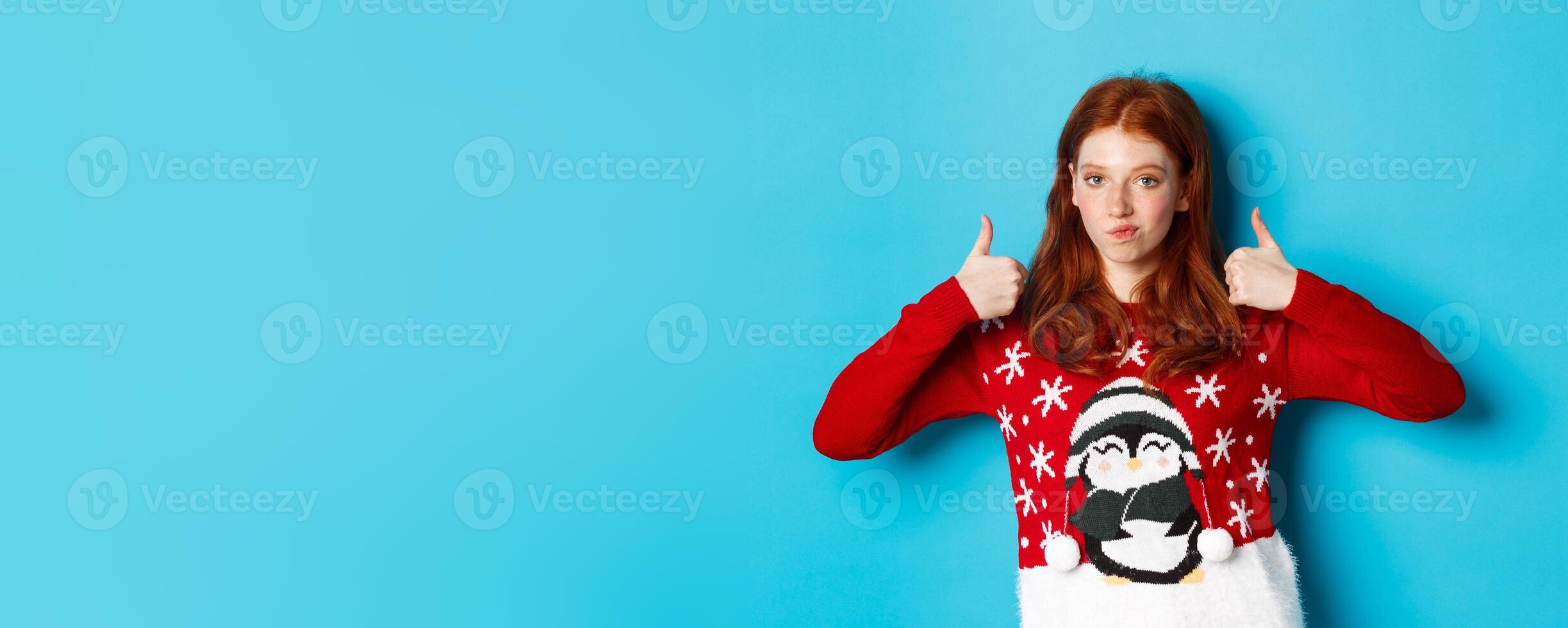 Winter holidays and Christmas Eve concept. Impressed redhead girl in xmas sweater, nod in approval and showing thumb up, praise good product, standing over blue background photo