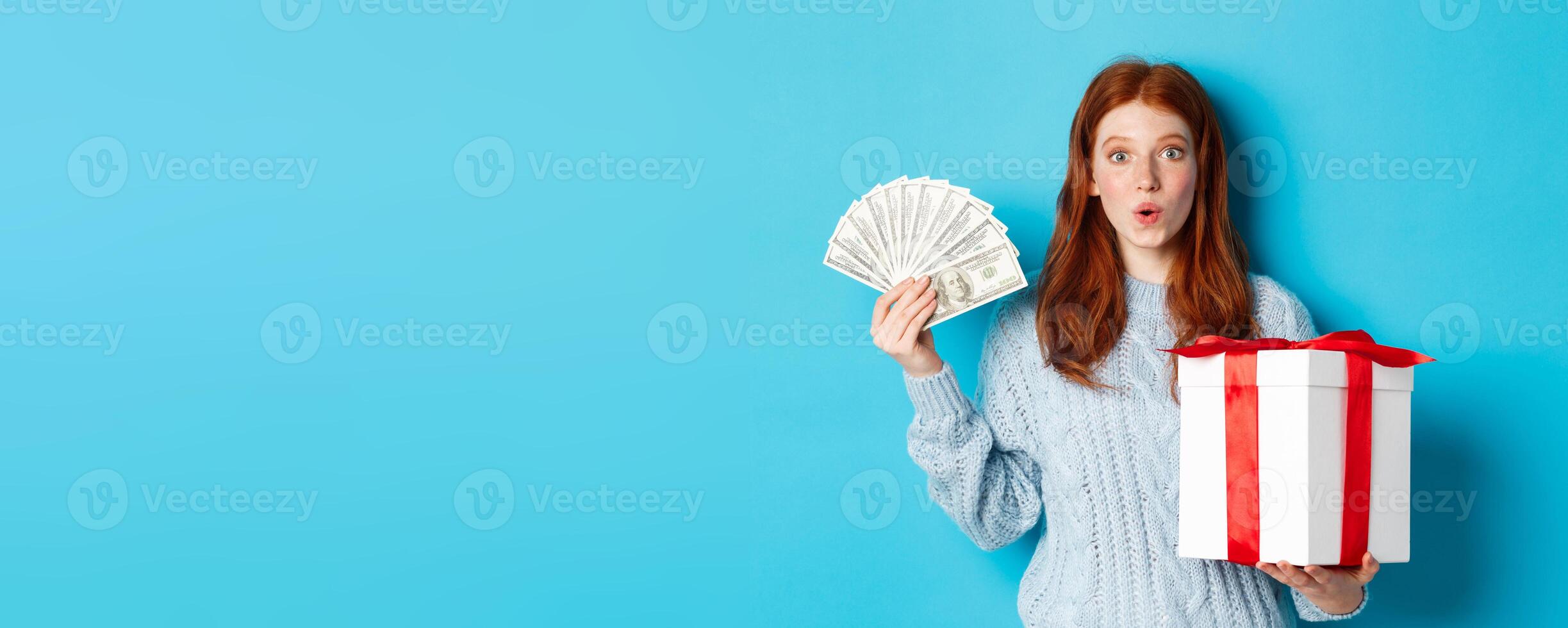 Christmas and shopping concept. Excited redhead girl looking at camera, holding big New Year gift and dollars, buying presents, standing over blue background photo