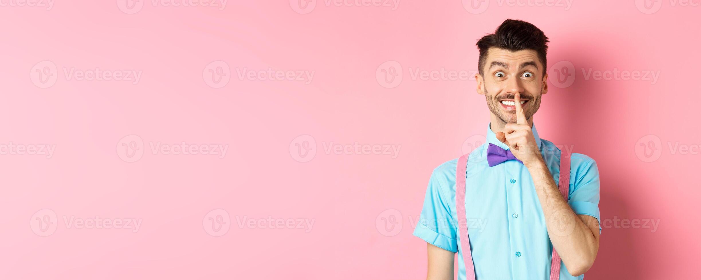 Cheerful young man making surprise, shushing at camera with happy smile, asking to keep voice down, be quiet, standing on pink background photo