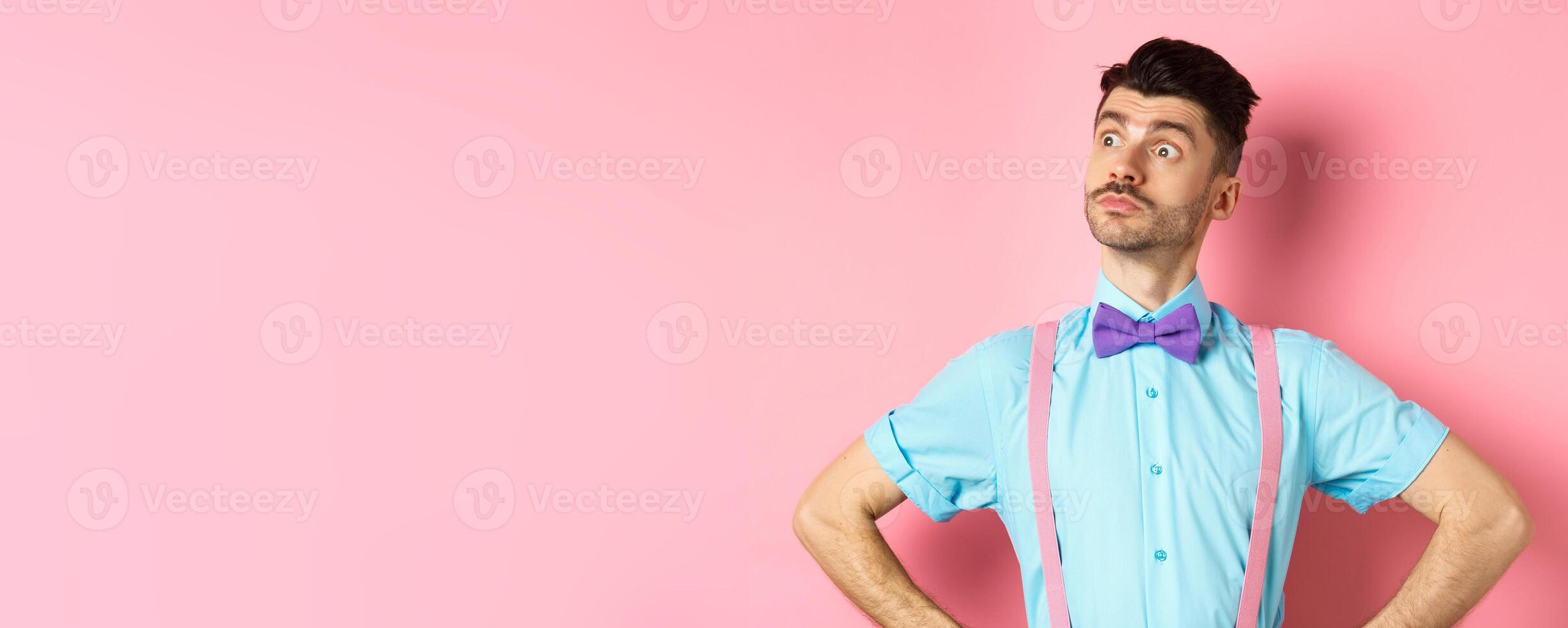 Funny caucasian man with moustache and bow-tie, standing confident with hands spread sideways, looking left at logo proud, standing on pink background photo