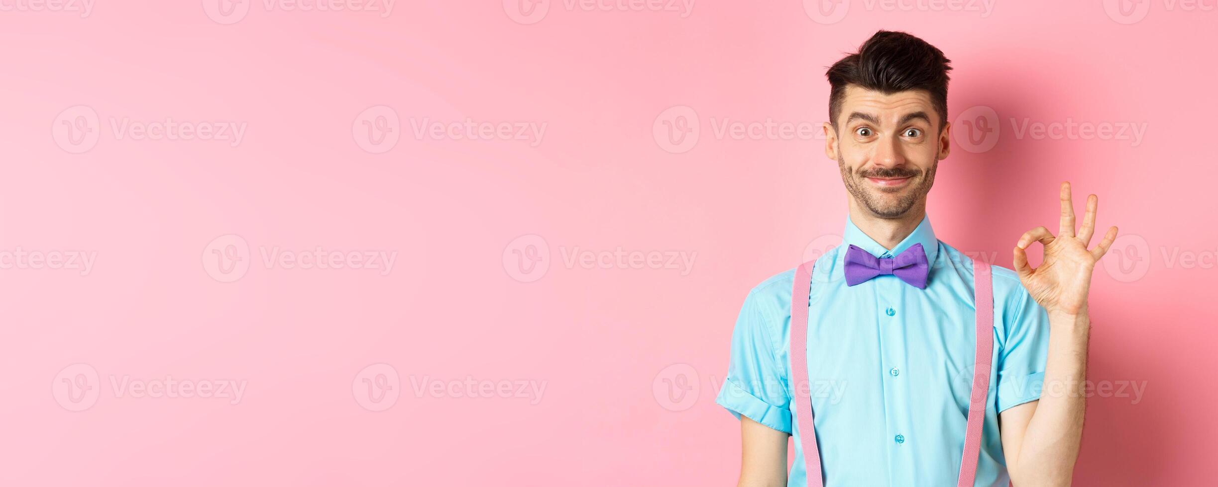 Smiling caucasian man in bow-tie and suspenders showing all okay gesture, recommending something cool, praising promo on pink background photo